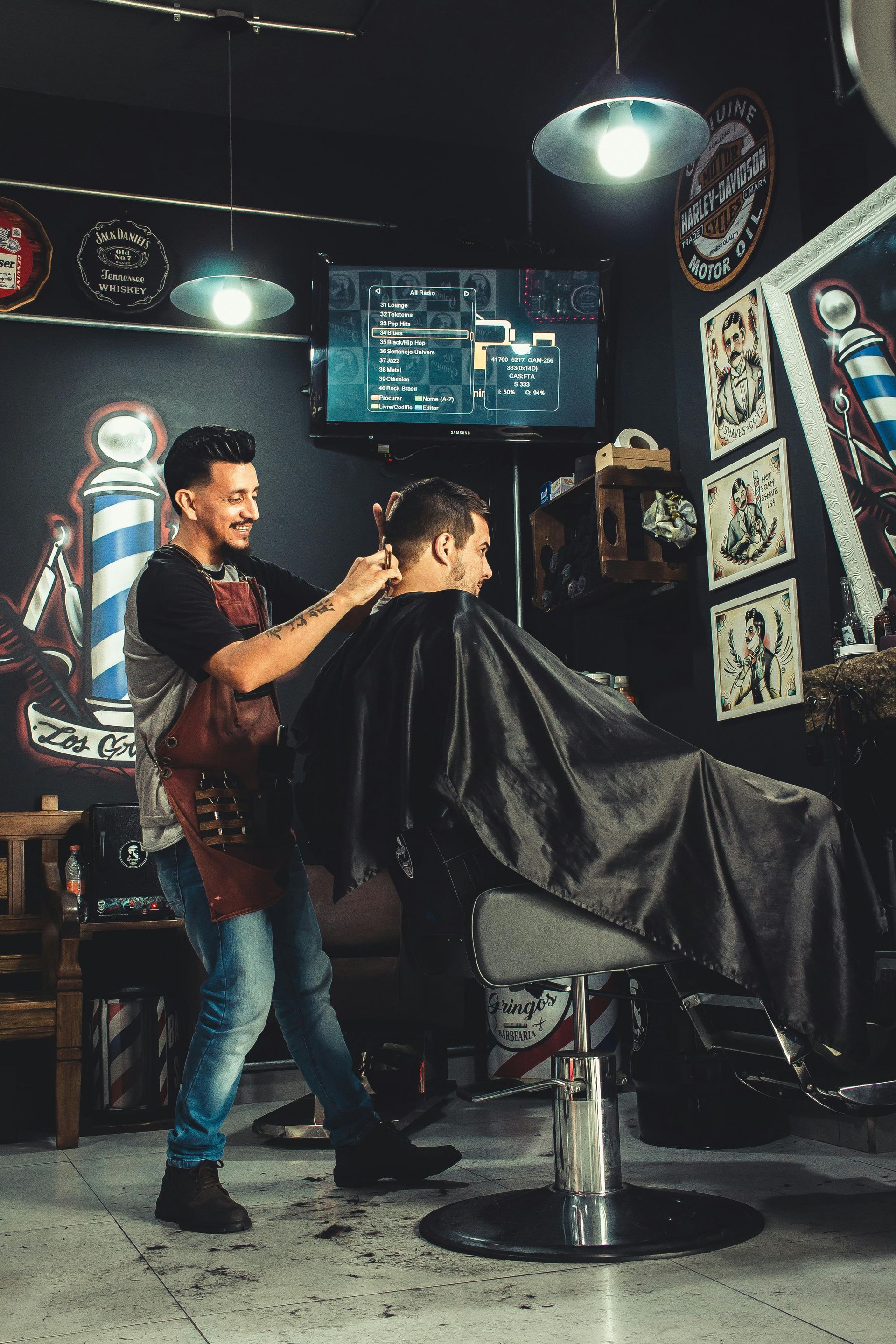 Barber cutting a client's hair in a dimly lit shop. Barber wears apron, client draped in cape.