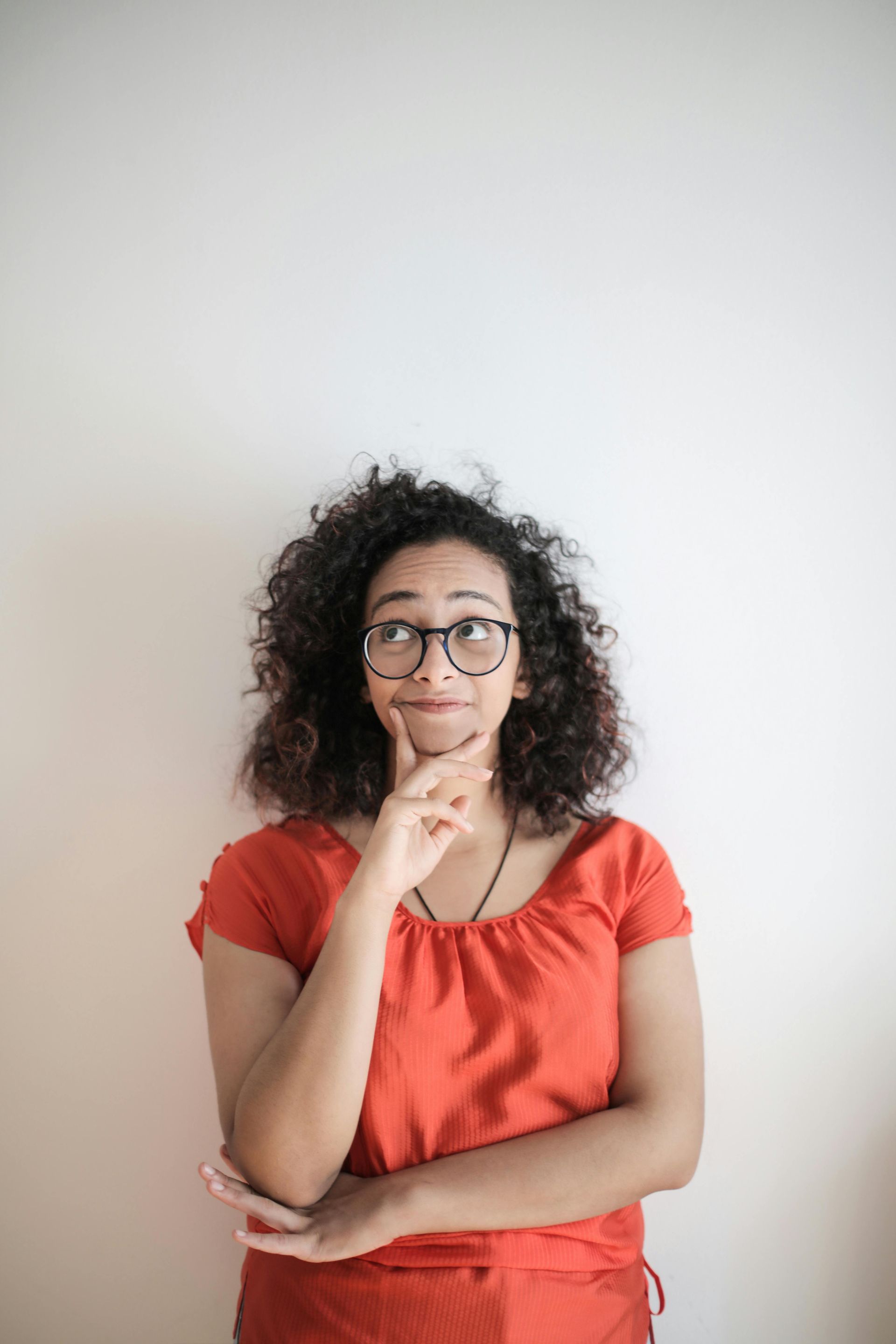 Woman in glasses and orange shirt, hand on chin, looking up thoughtfully against a white wall.