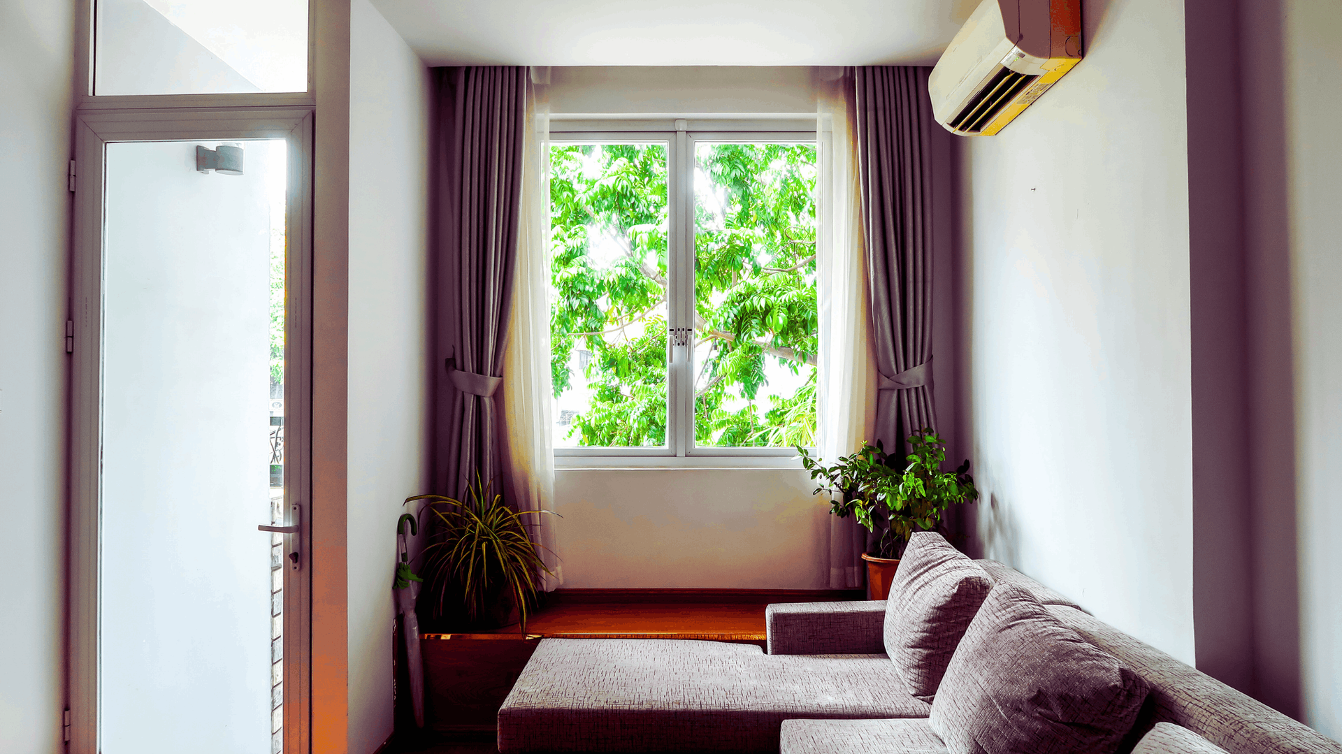 Living room with a window showing trees, gray curtains, and a sofa. An air conditioner is mounted on the wall — Chayser Electrical in Currimundi, QLD