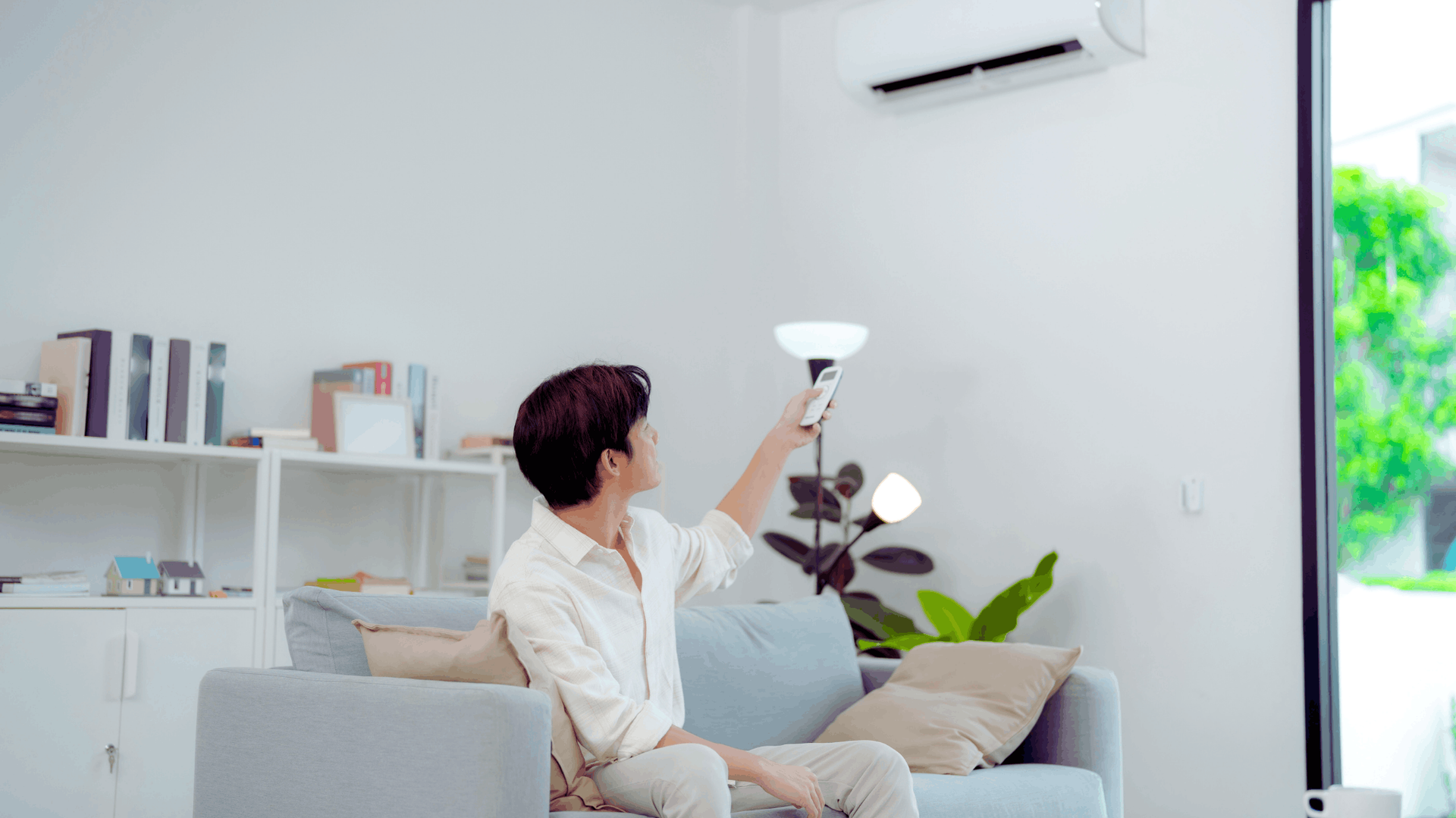 Person sitting on couch adjusting air conditioner with remote. White room with bookshelf — Chayser Electrical in Currimundi, QLD