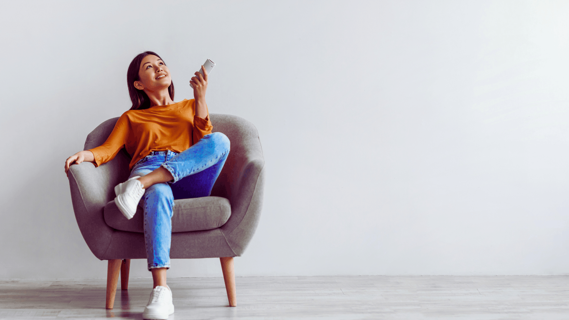 Woman seated in a gray armchair, looking up, holding a phone. She wears orange top, blue jeans, white sneakers. White wall background — Chayser Electrical in Currimundi, QLD