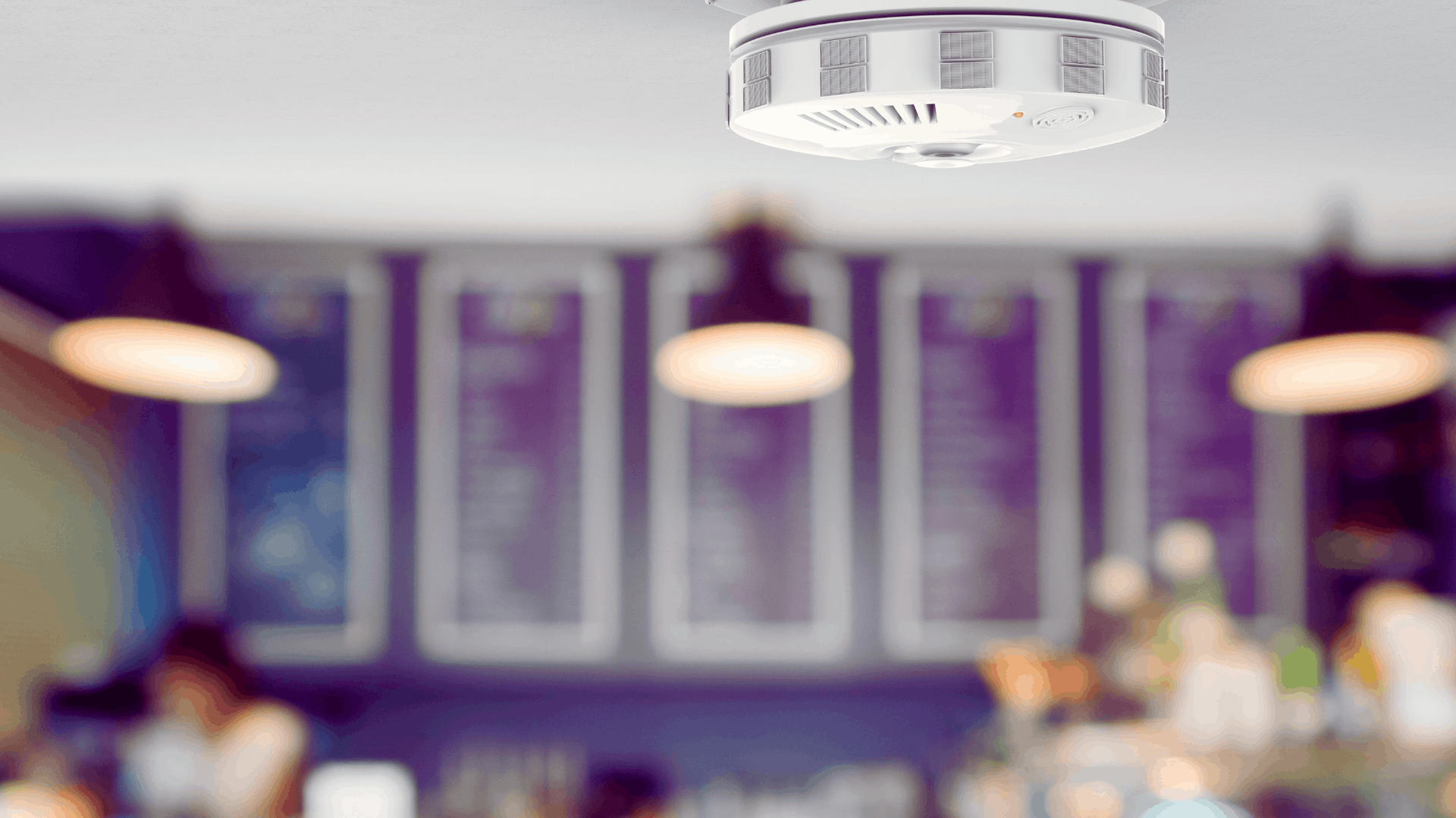 Smoke detector mounted on a ceiling, blurred cafe background with pendant lights — Chayser Electrical in Currimundi, QLD
