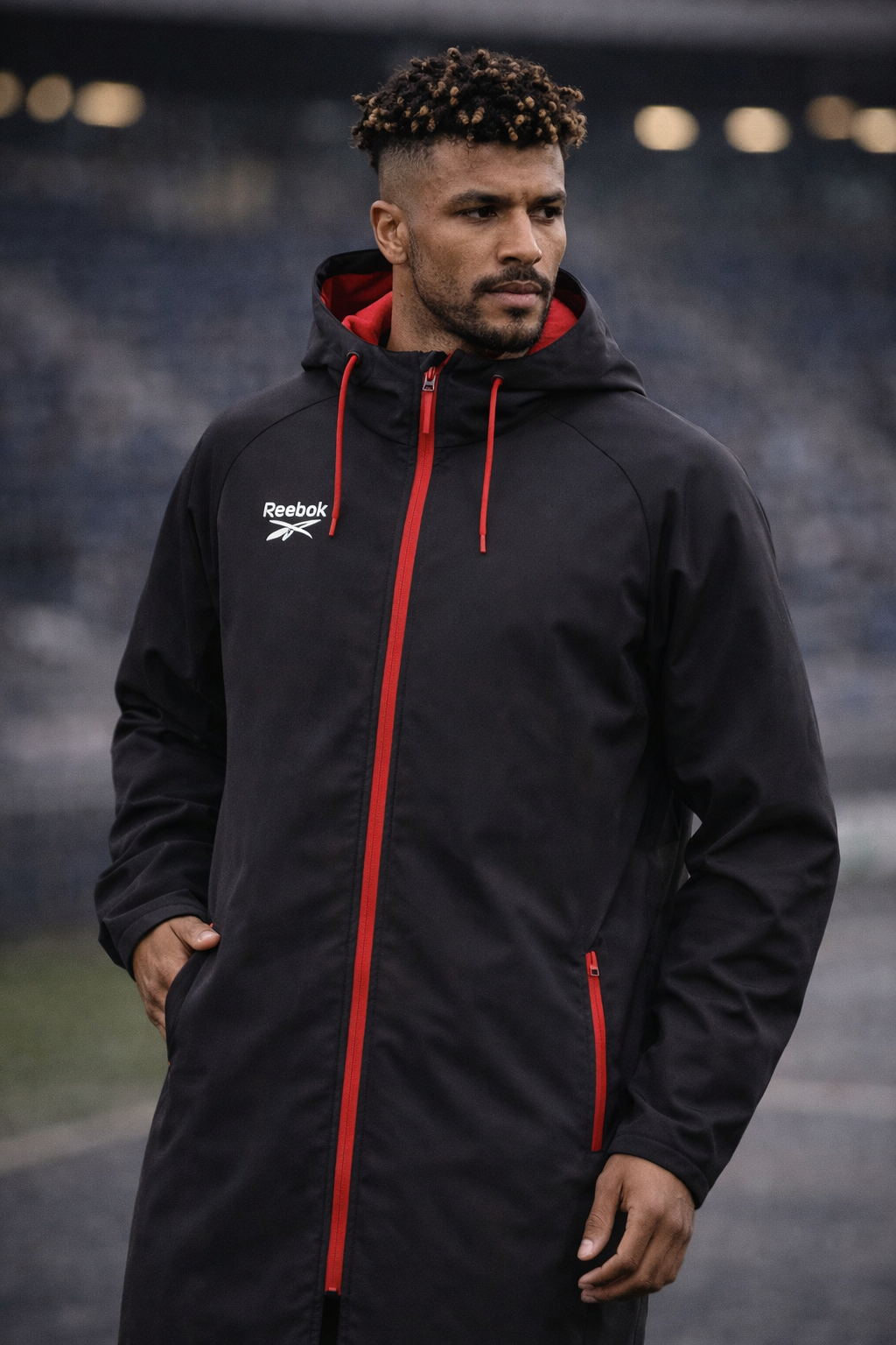 Soccer player in black hooded jacket with red trim standing on a stadium field