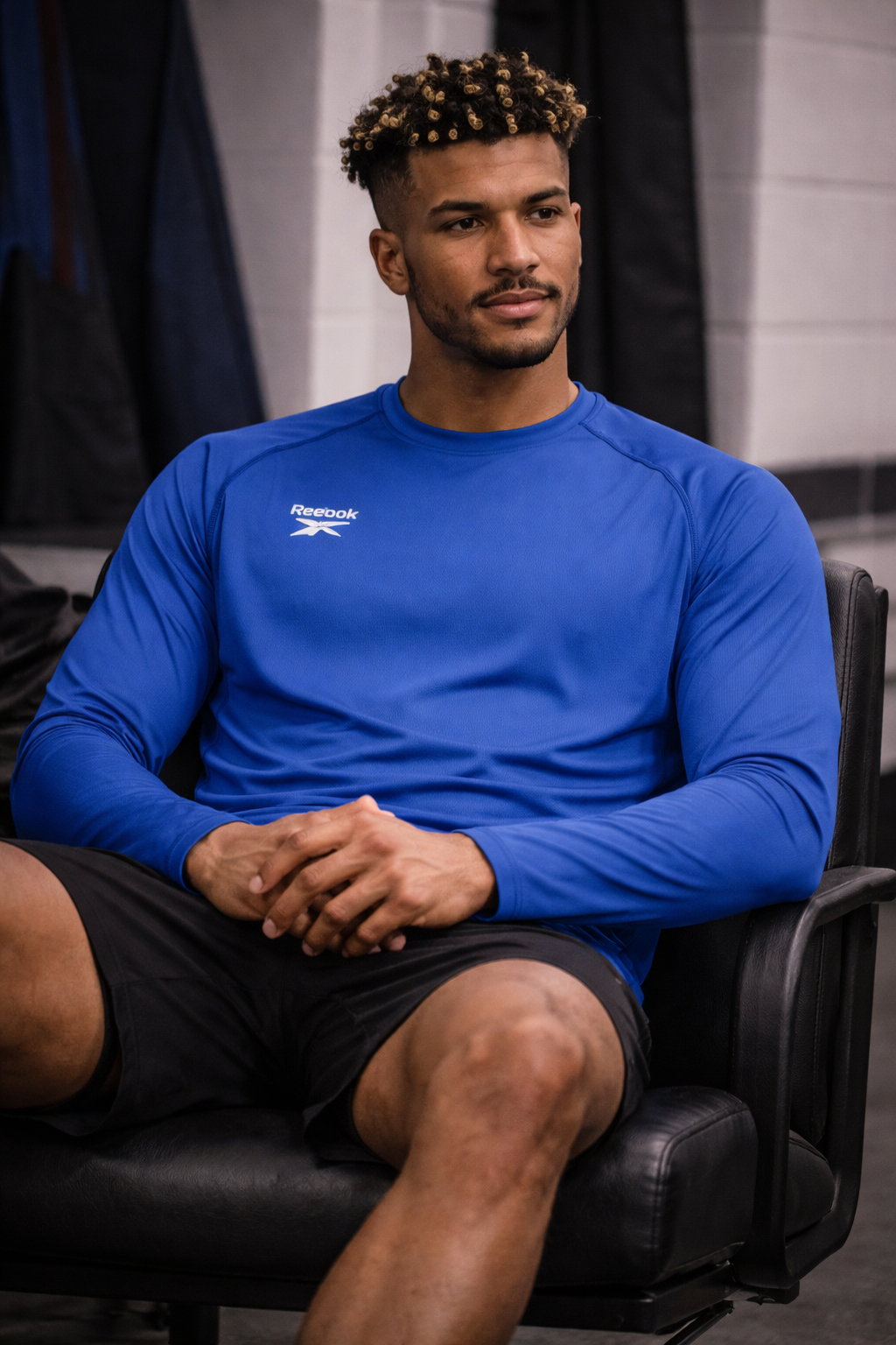 Man in blue long-sleeve shirt and black shorts sitting in a chair, looking off to the side.