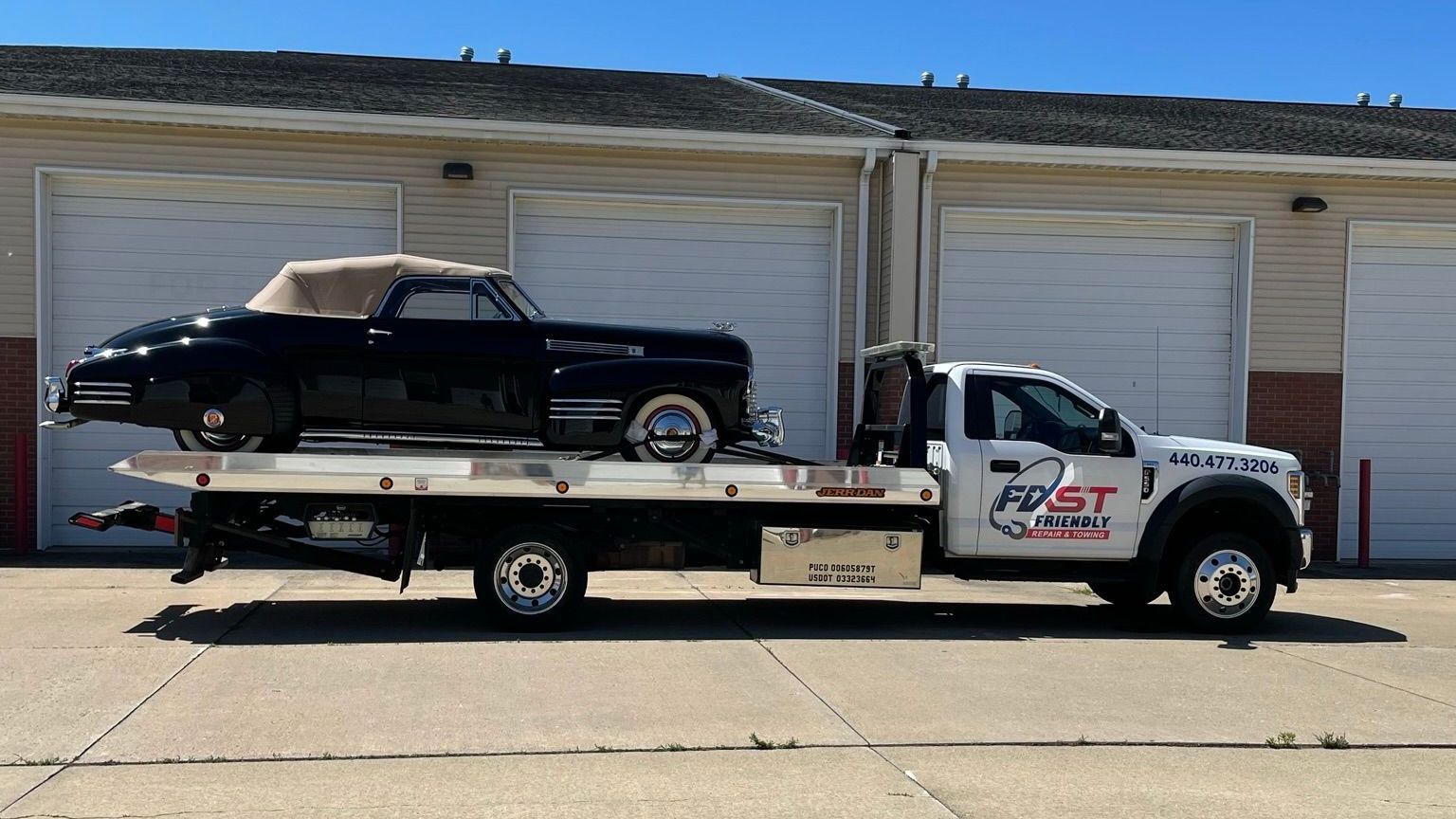A tow truck with a black car on the back is parked in front of a garage.