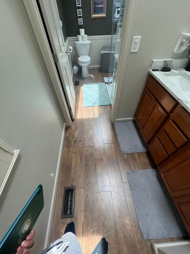 Narrow hallway with wood floor leading to a small bathroom with toilet and blue bath mat.