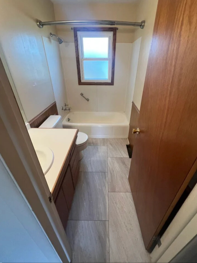 Narrow bathroom with a tub, toilet, sink, wood door, and a small window