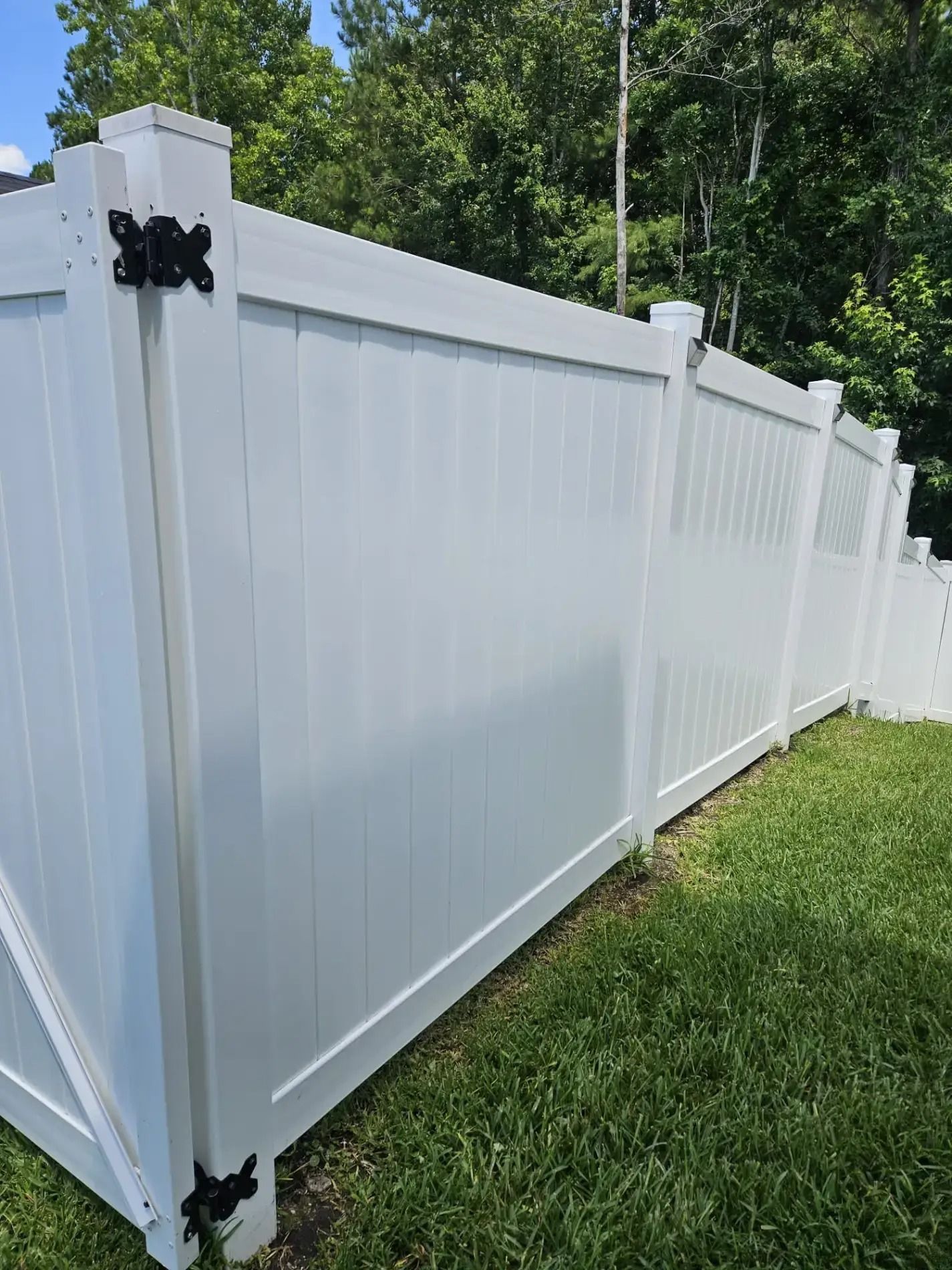 White vinyl privacy fence with black hinges along a grassy yard, viewed at an angle