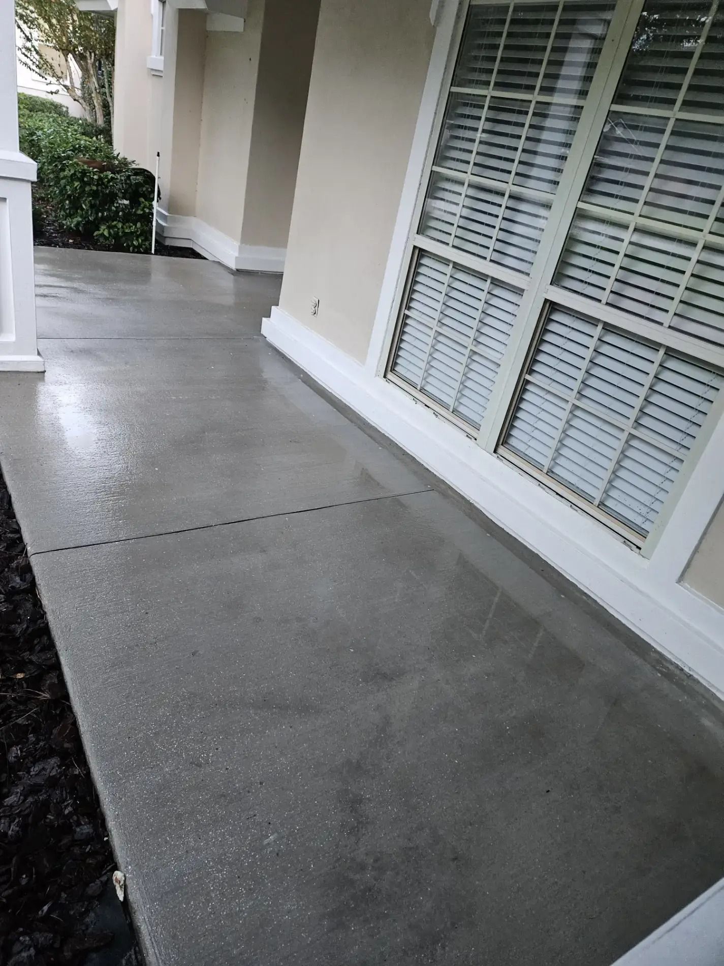 Wet concrete patio beside beige house with white-trimmed windows and columns