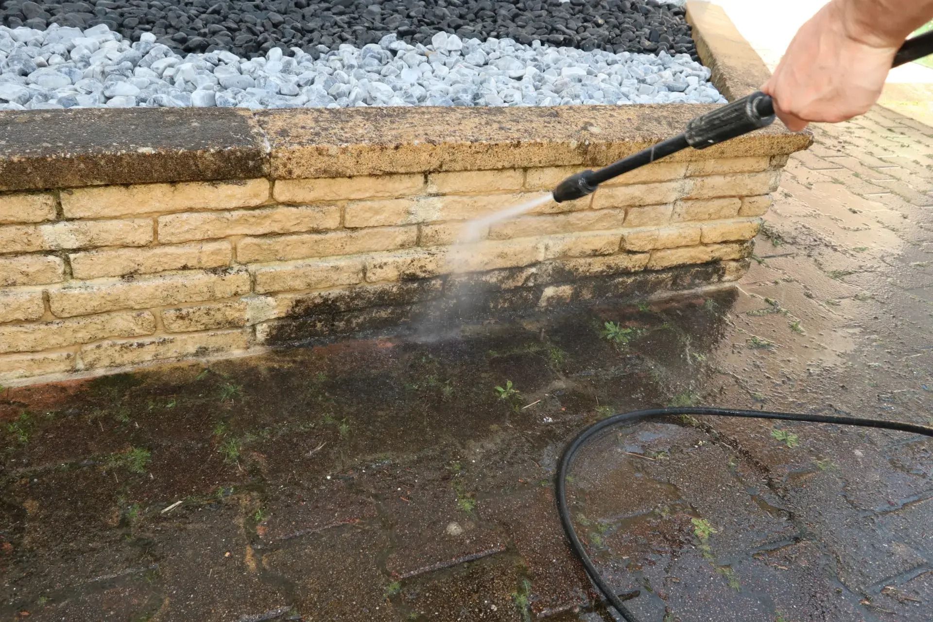 Person pressure-washing a brick wall beside gravel and wet pavement