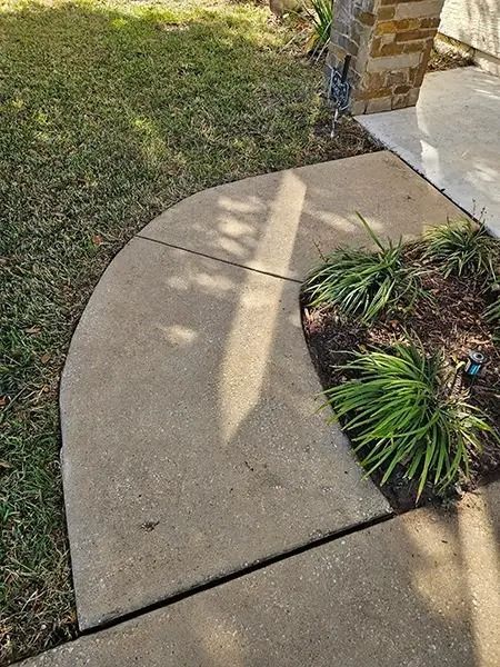 Curved concrete sidewalk beside grass and a small landscaped plant bed near a brick porch pillar