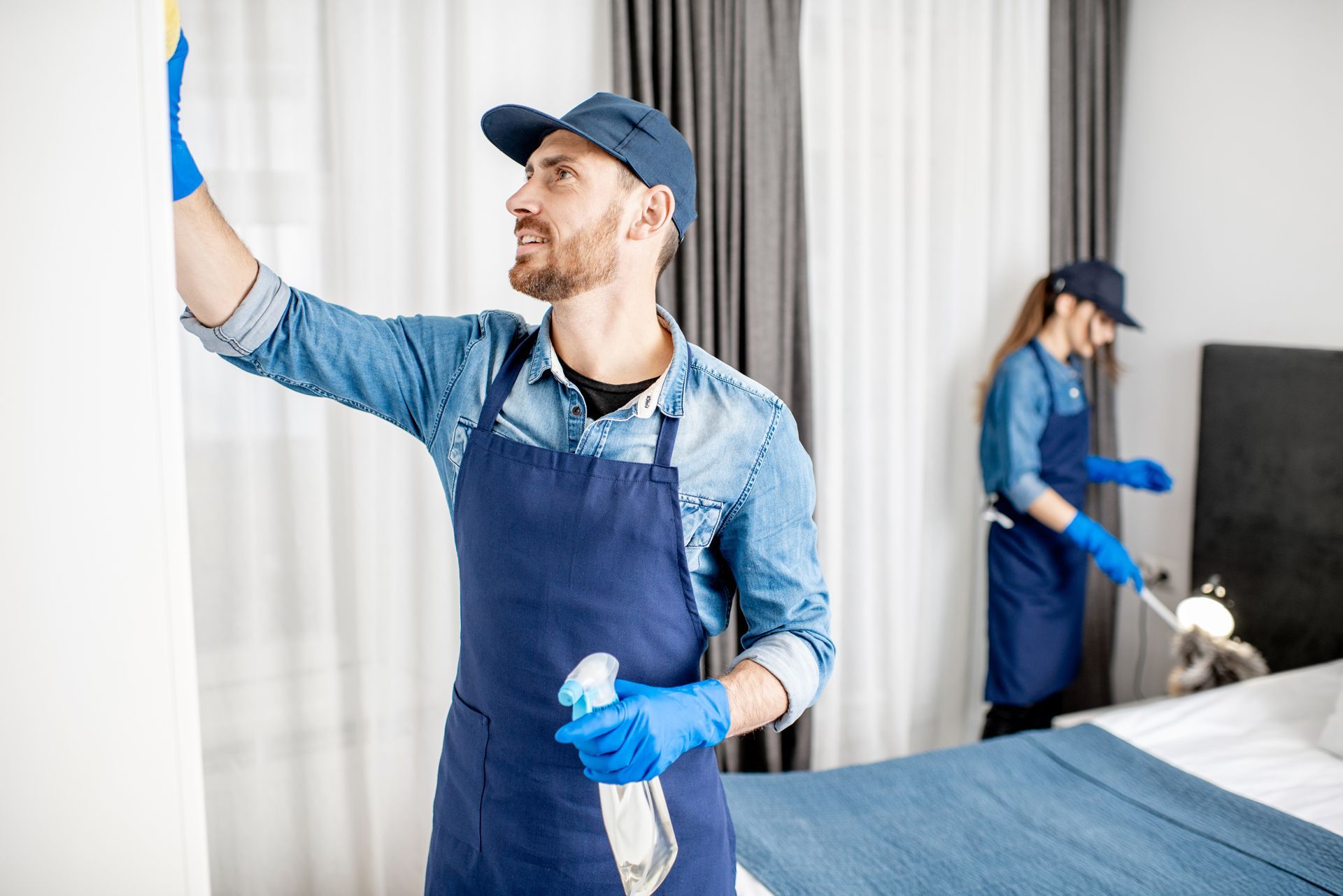 Woman wiping kitchen cabinet with pink cloth while man washes dishes in the background