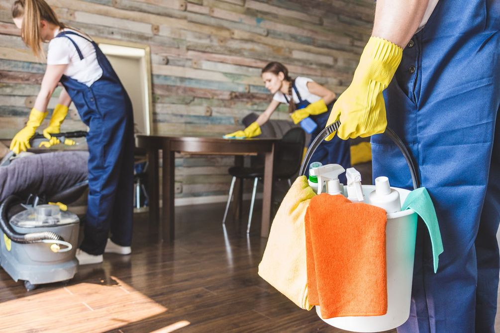 People cleaning a wooden floor with mops and supplies in a room with a stone wall