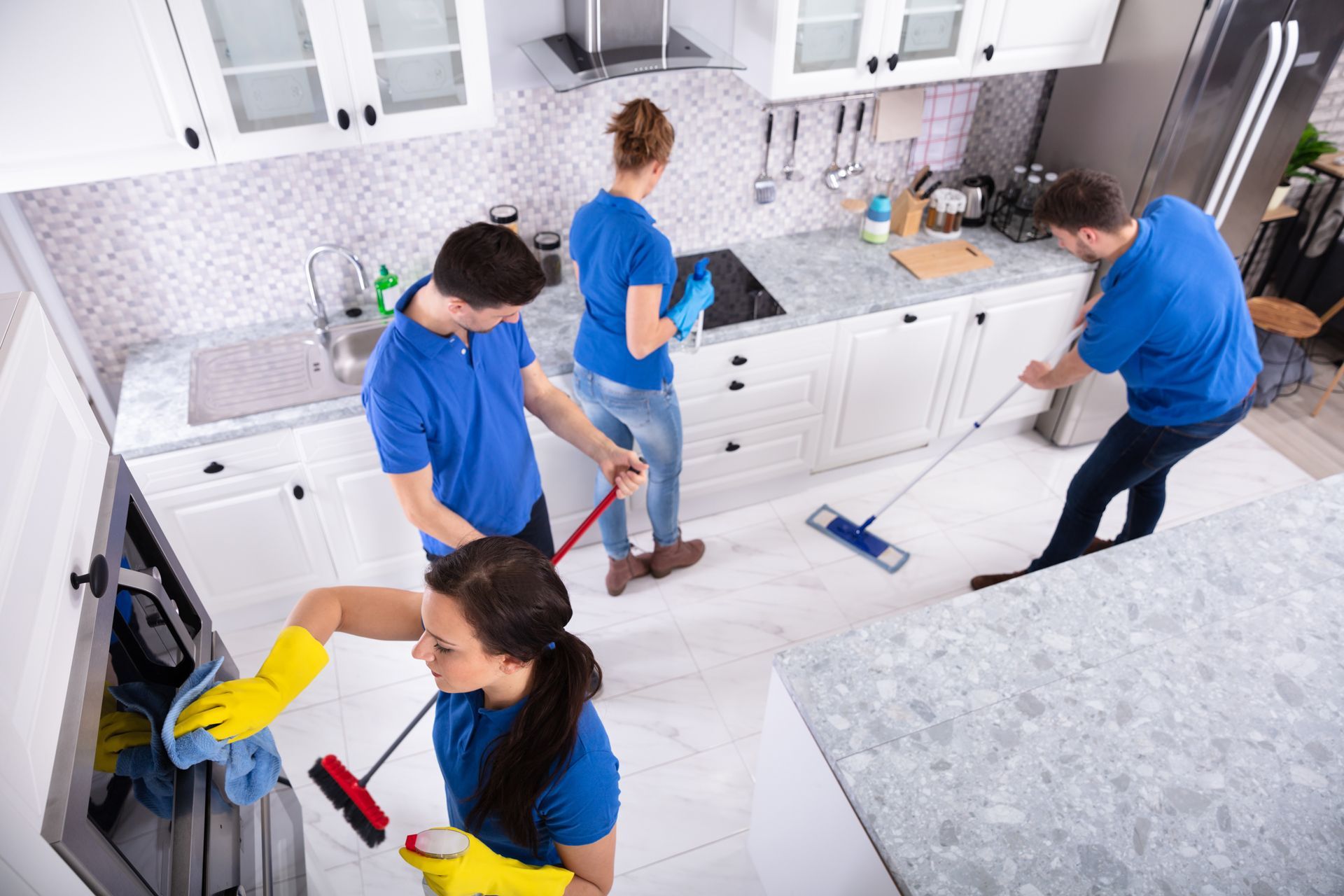 People cleaning a bright kitchen with brooms, mops, and spray bottles