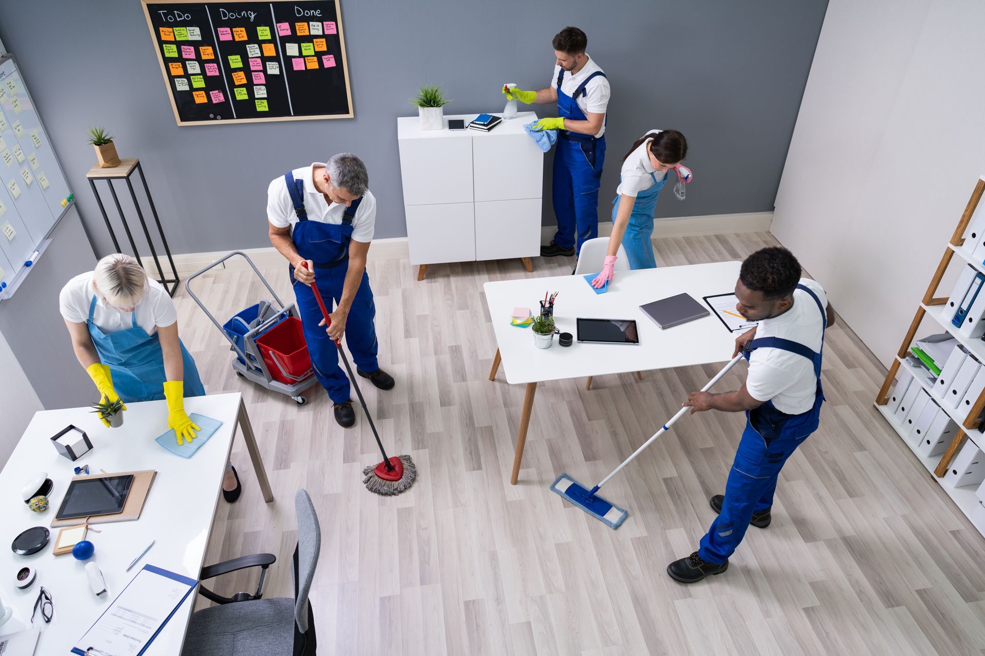 Several workers in blue overalls clean a bright office with mops and buckets