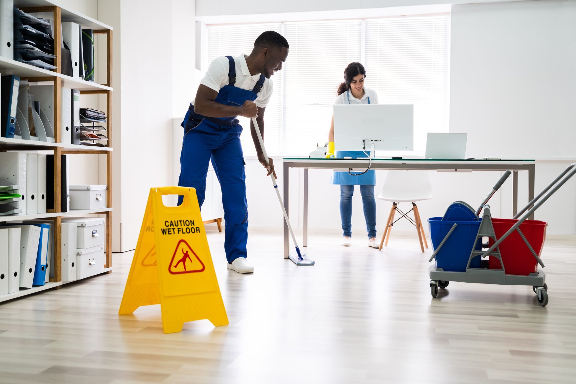 Office cleaners mopping a bright workspace with a yellow wet floor sign and cleaning cart