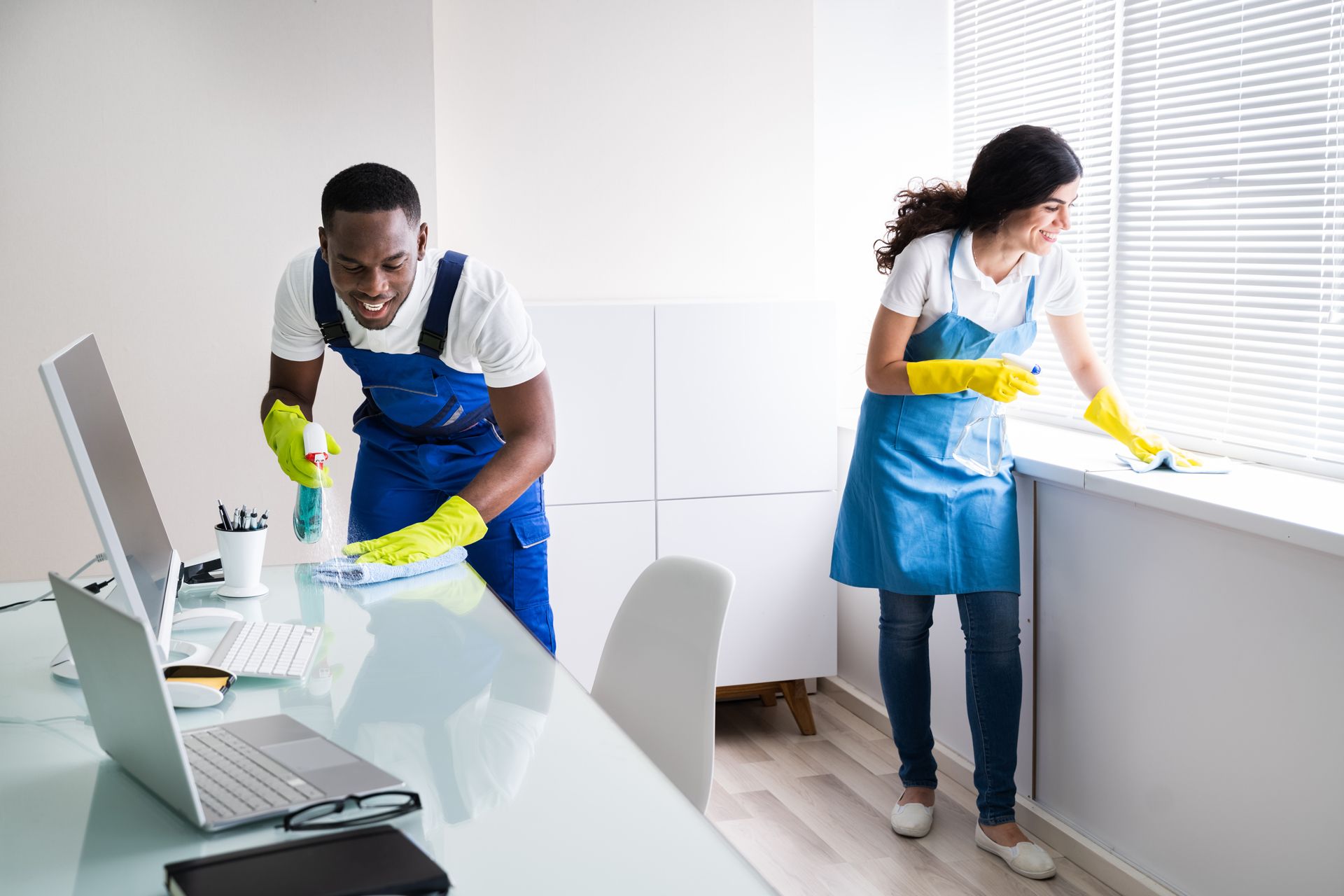 Two cleaners in blue aprons and gloves disinfecting a bright office with a desk and laptop
