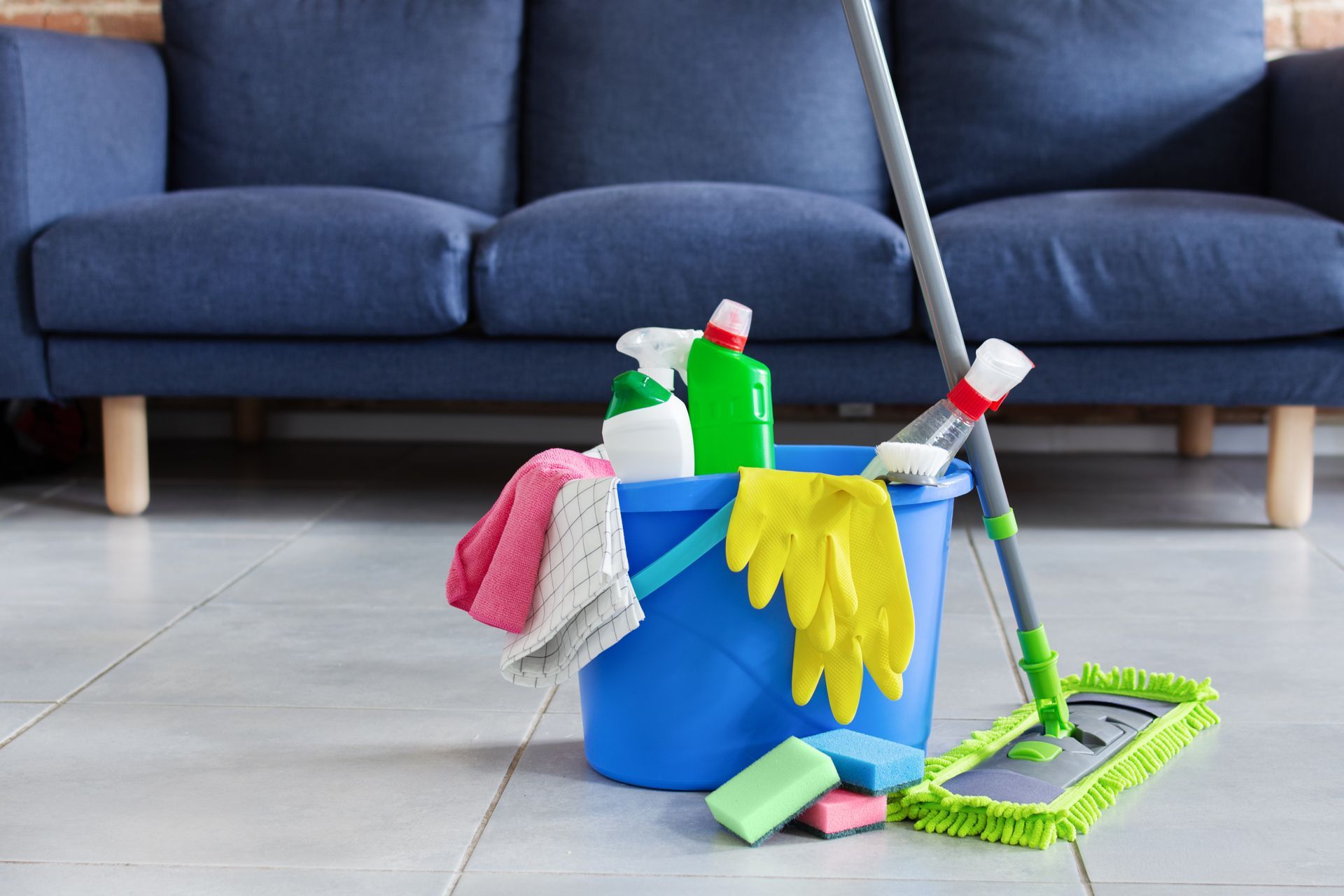 Cleaning supplies in a blue bucket beside a mop and broom on a tiled floor in front of a couch