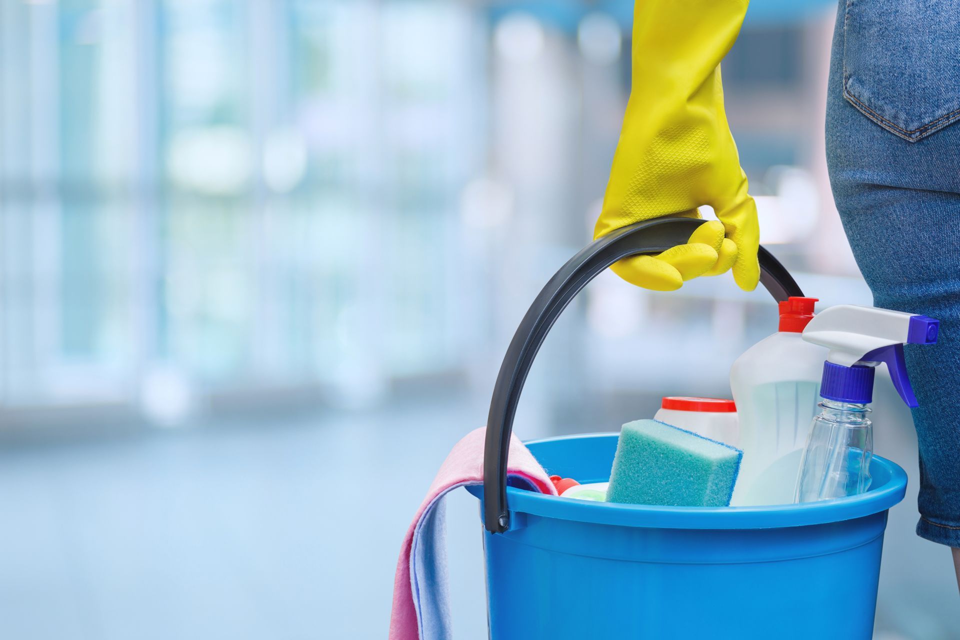 Gloved hand holding a spray bottle beside a blue cleaning bucket with supplies