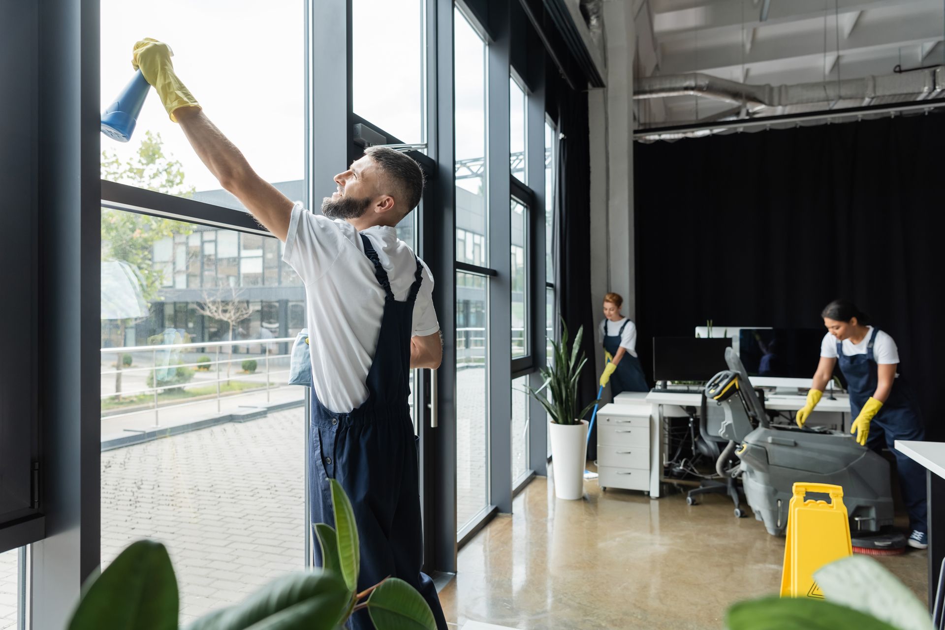 Person cleaning tall windows in a bright office while another cleans equipment in the background.
