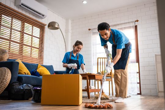 Two people cleaning a living room, with one mopping the floor and the other dusting near a box.