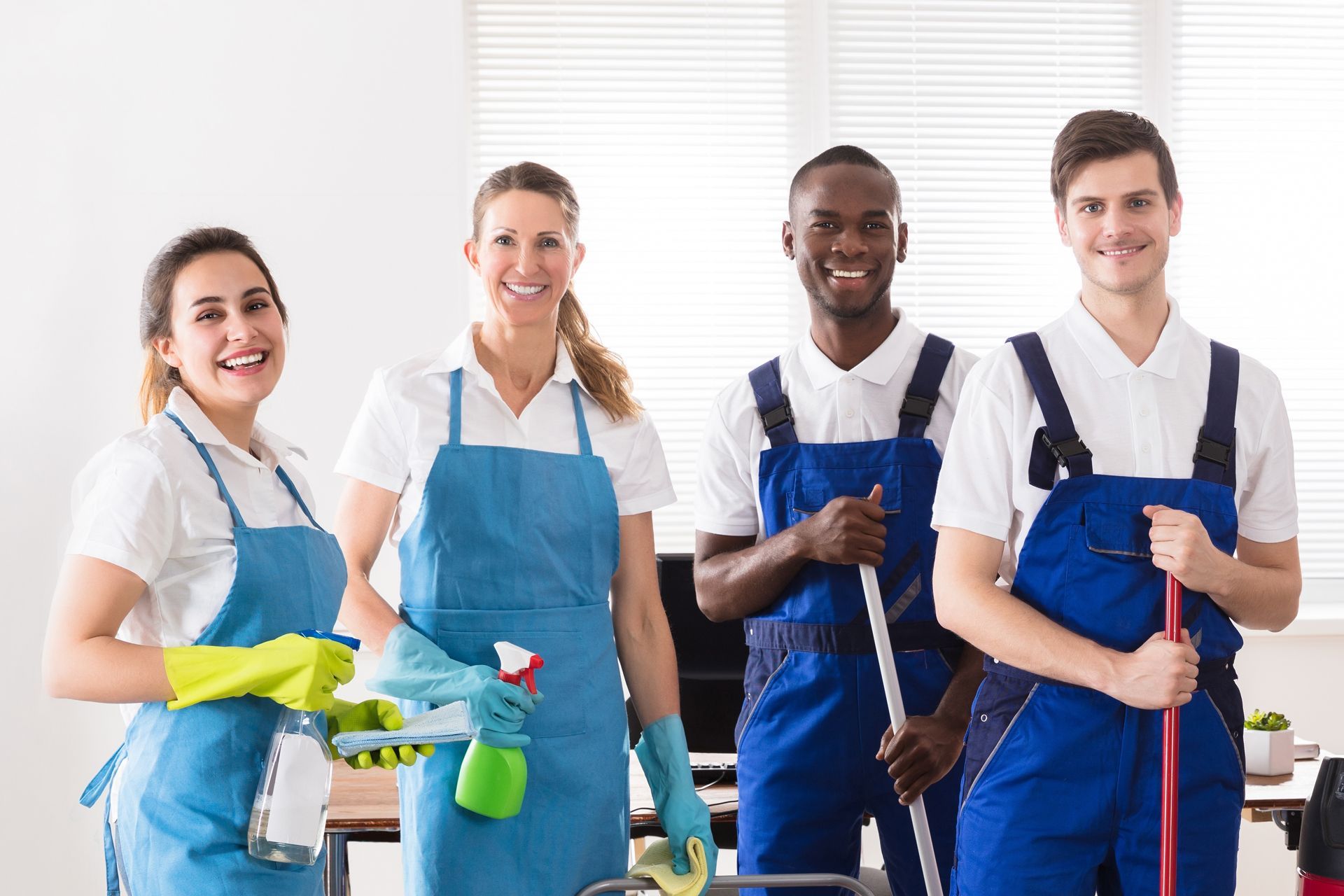 Four cleaners in blue overalls stand with cleaning supplies in a bright room