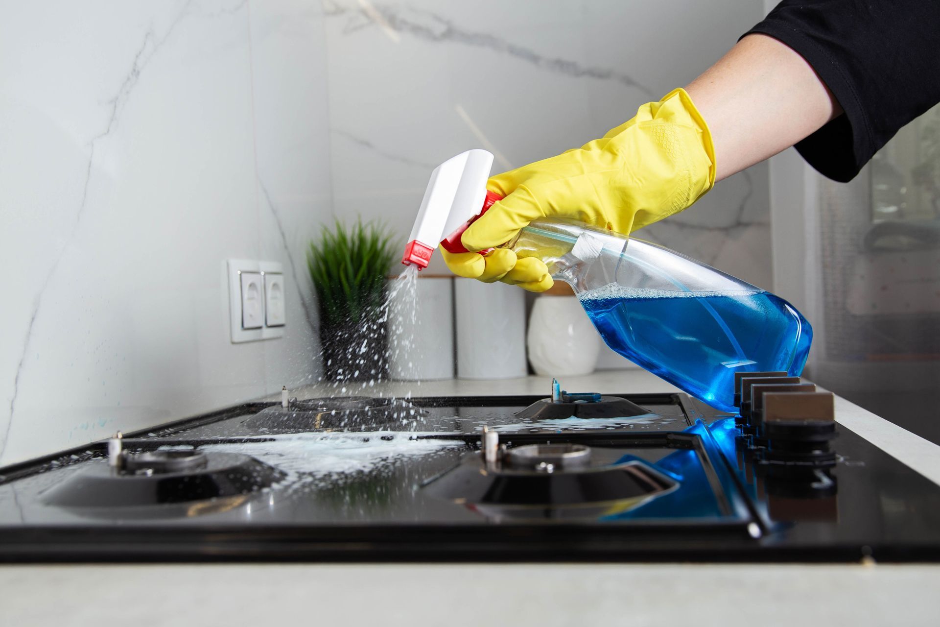 Gloved hand spraying blue cleaner onto a stovetop with soap suds in a kitchen
