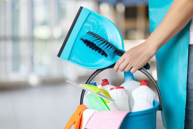Person holding a blue cleaning caddy with sprays, cloths, and a dustpan with broom.