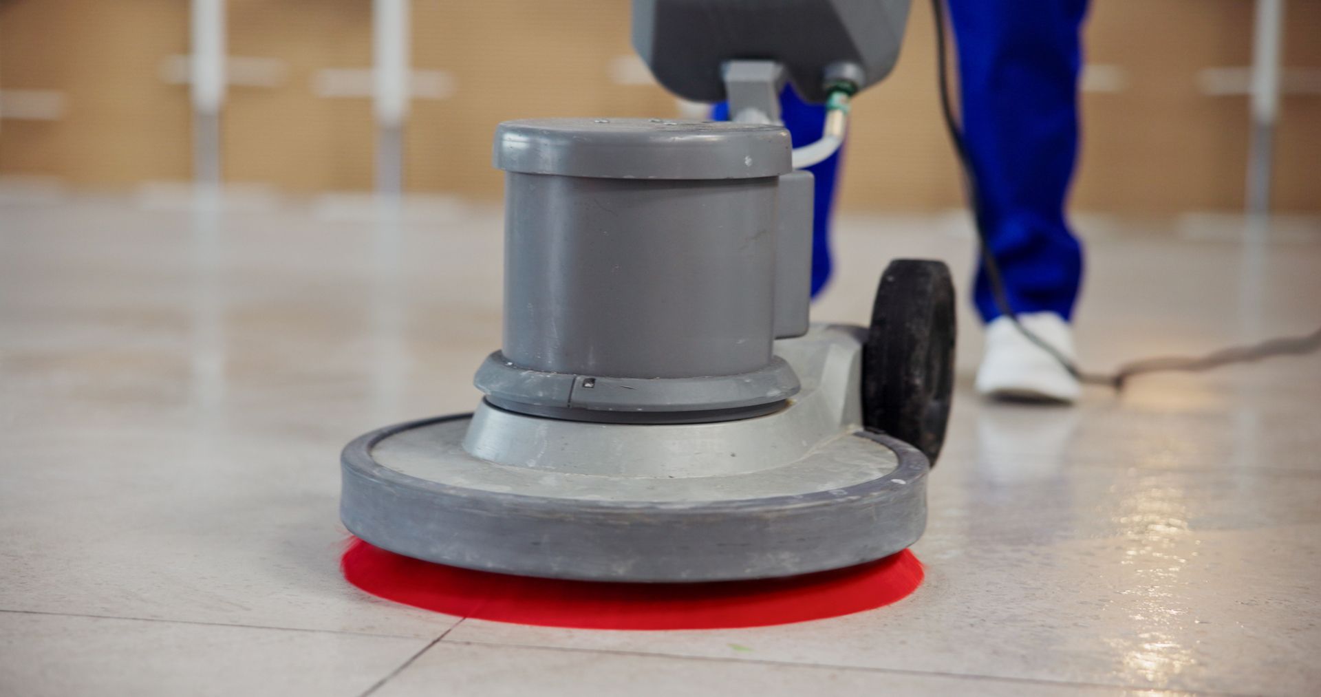 Floor buffer cleaning a tile floor in a commercial space, with a red pad and a person in blue pants behind it