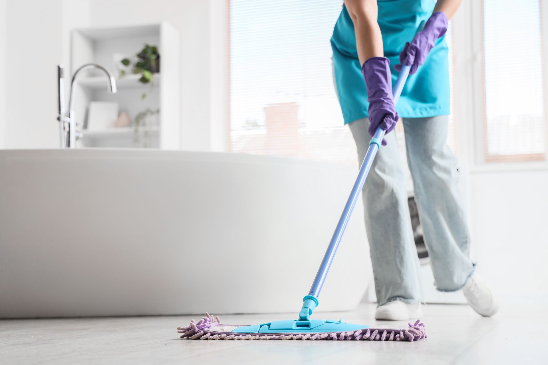 Person mopping a bright bathroom floor with a purple mop and gloves