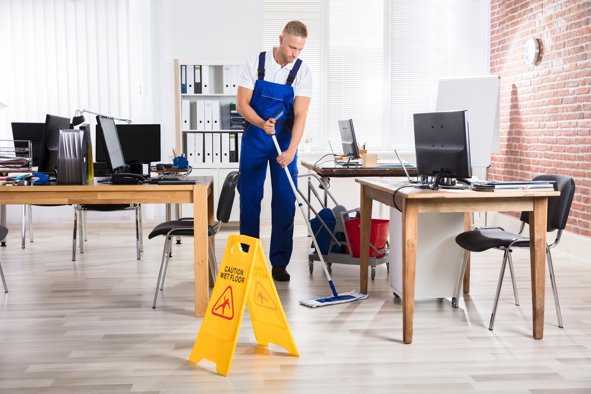 Office cleaner mopping floor beside a yellow wet-floor sign in a bright workspace