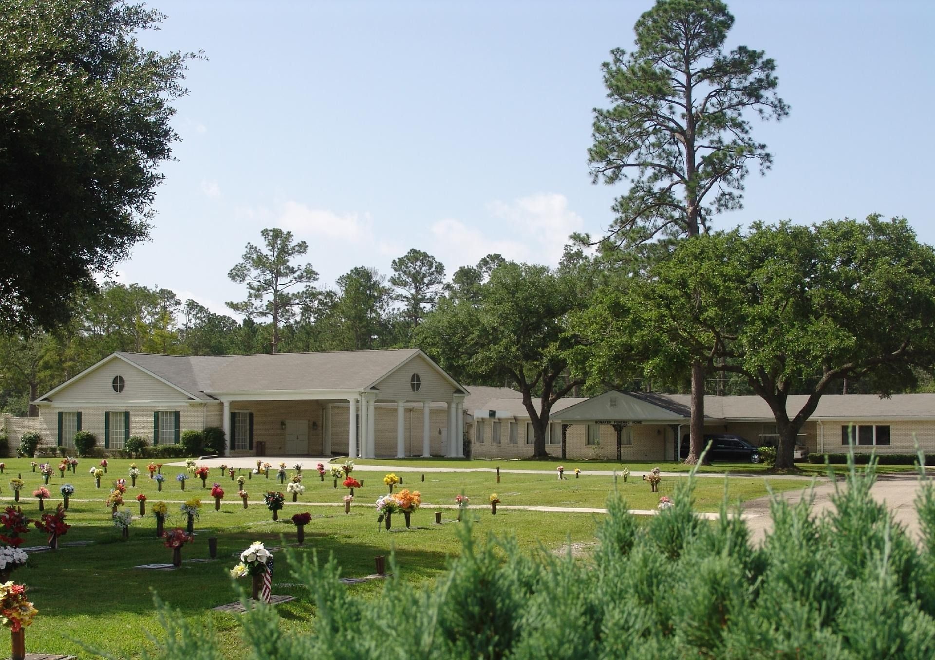 Garden with rows of small flower crosses in front of a light-colored building and tall pine trees