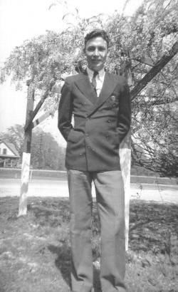 Man in a suit standing outdoors beside a tree, with a road and buildings in the background.