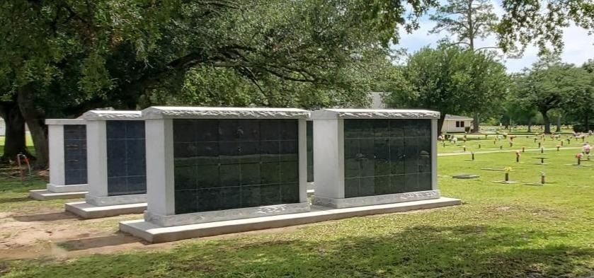 Three gray columbarium walls in a grassy cemetery with trees and headstones