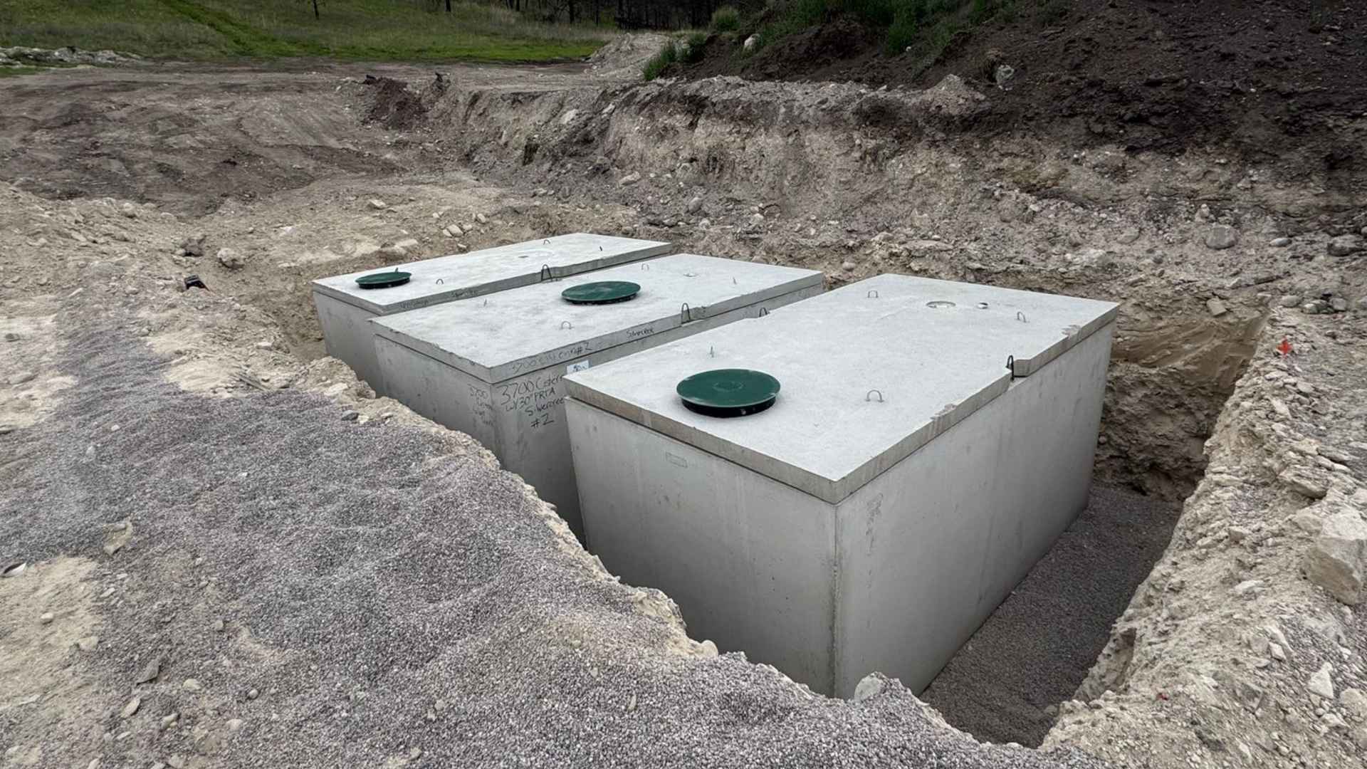 Three concrete septic tanks in a dirt excavation pit, with a cloudy sky and wooded hill in the background.