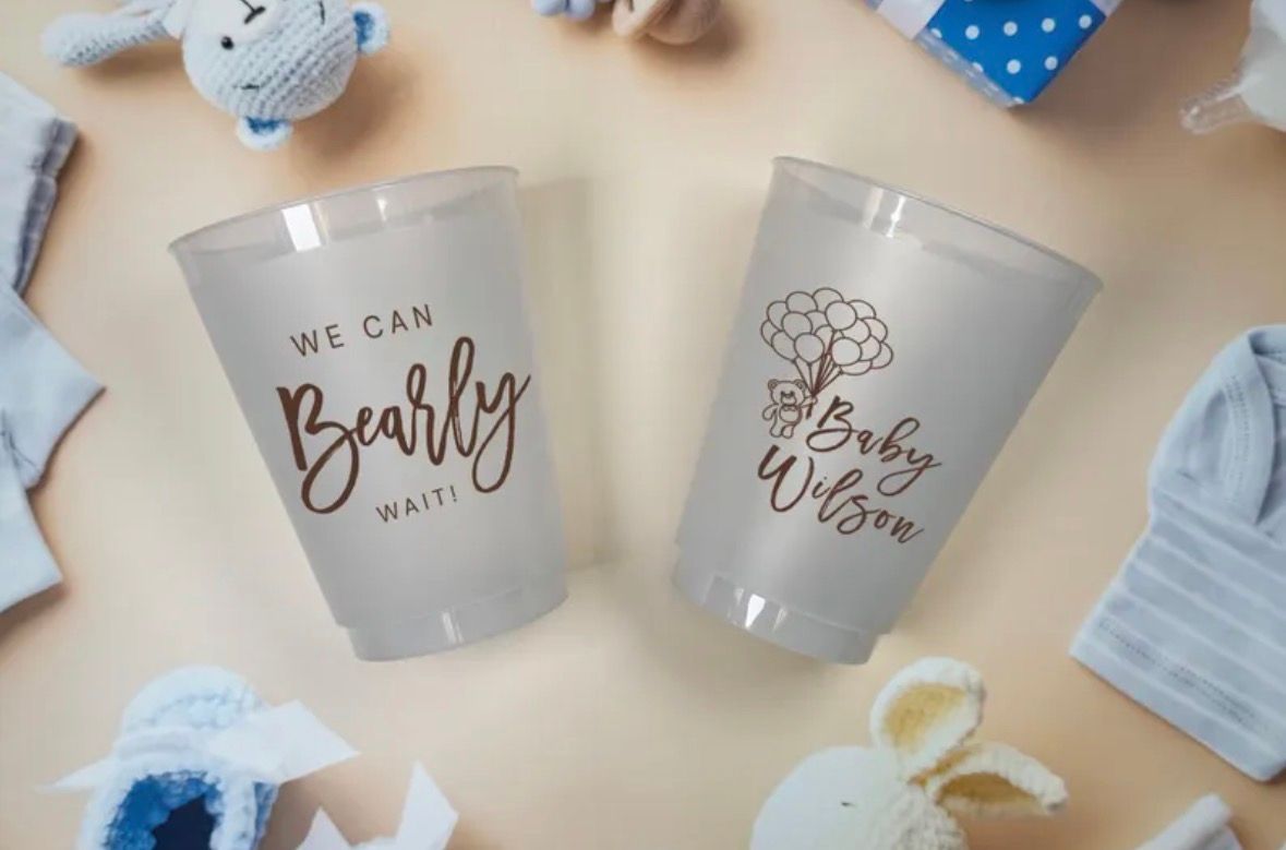 Two frosted plastic cups with baby shower text, surrounded by baby items.