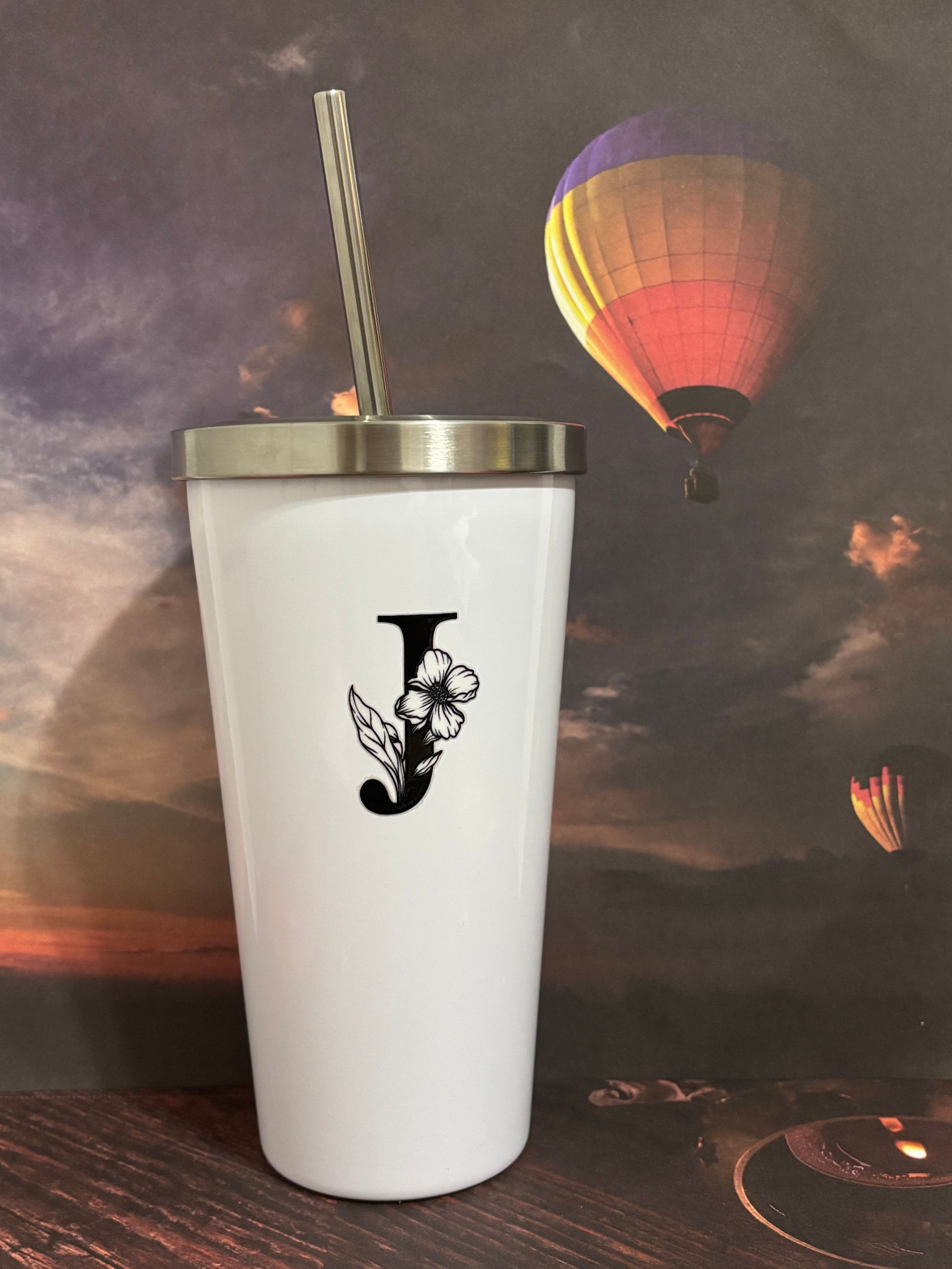White tumbler with black logo and metal straw in front of a hot air balloon backdrop
