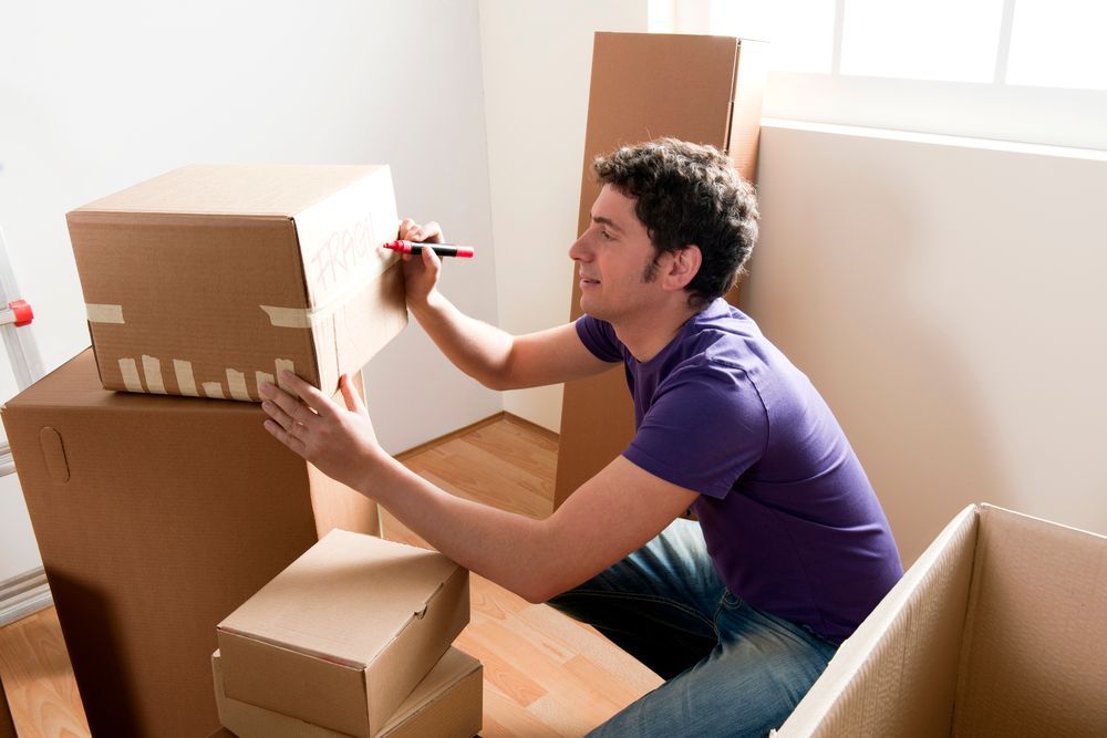 Person labeling a cardboard box with a red marker in a sunlit room surrounded by moving boxes