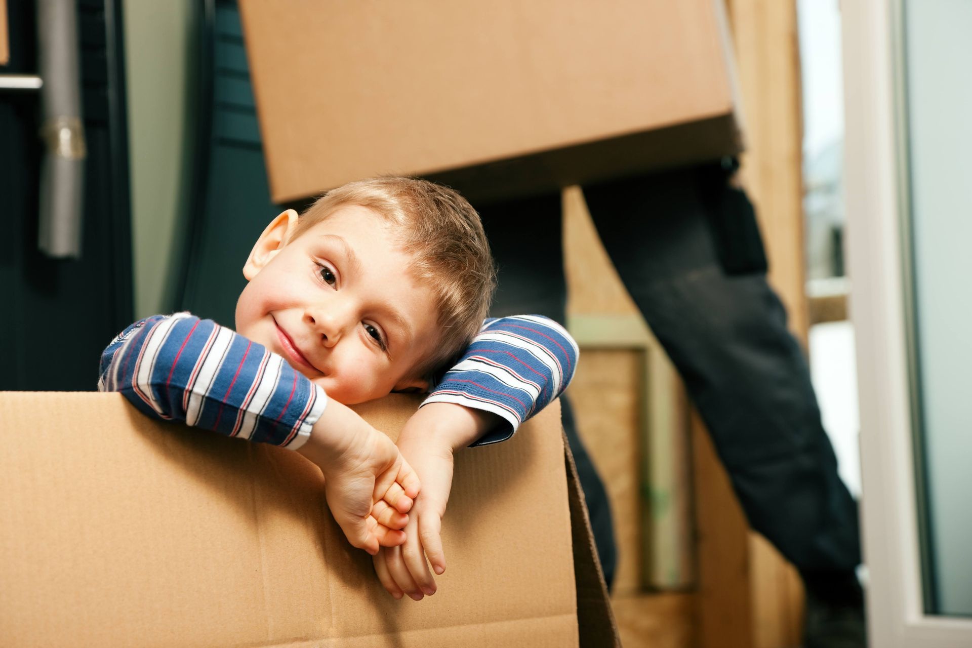 Child smiling from inside a cardboard box, with arms resting on the edge in a room.