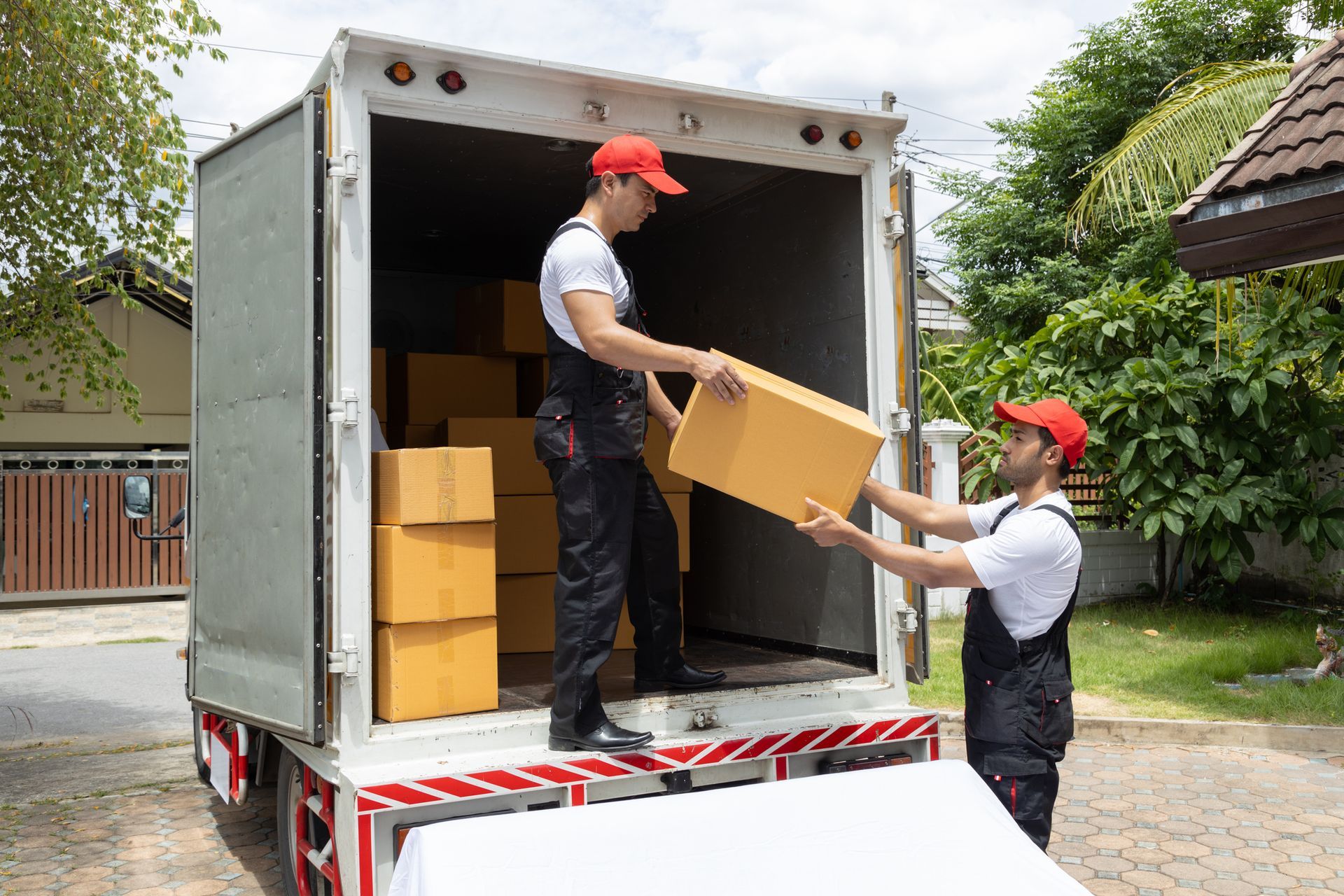 Two movers in red caps loading cardboard boxes into a white moving truck outdoors