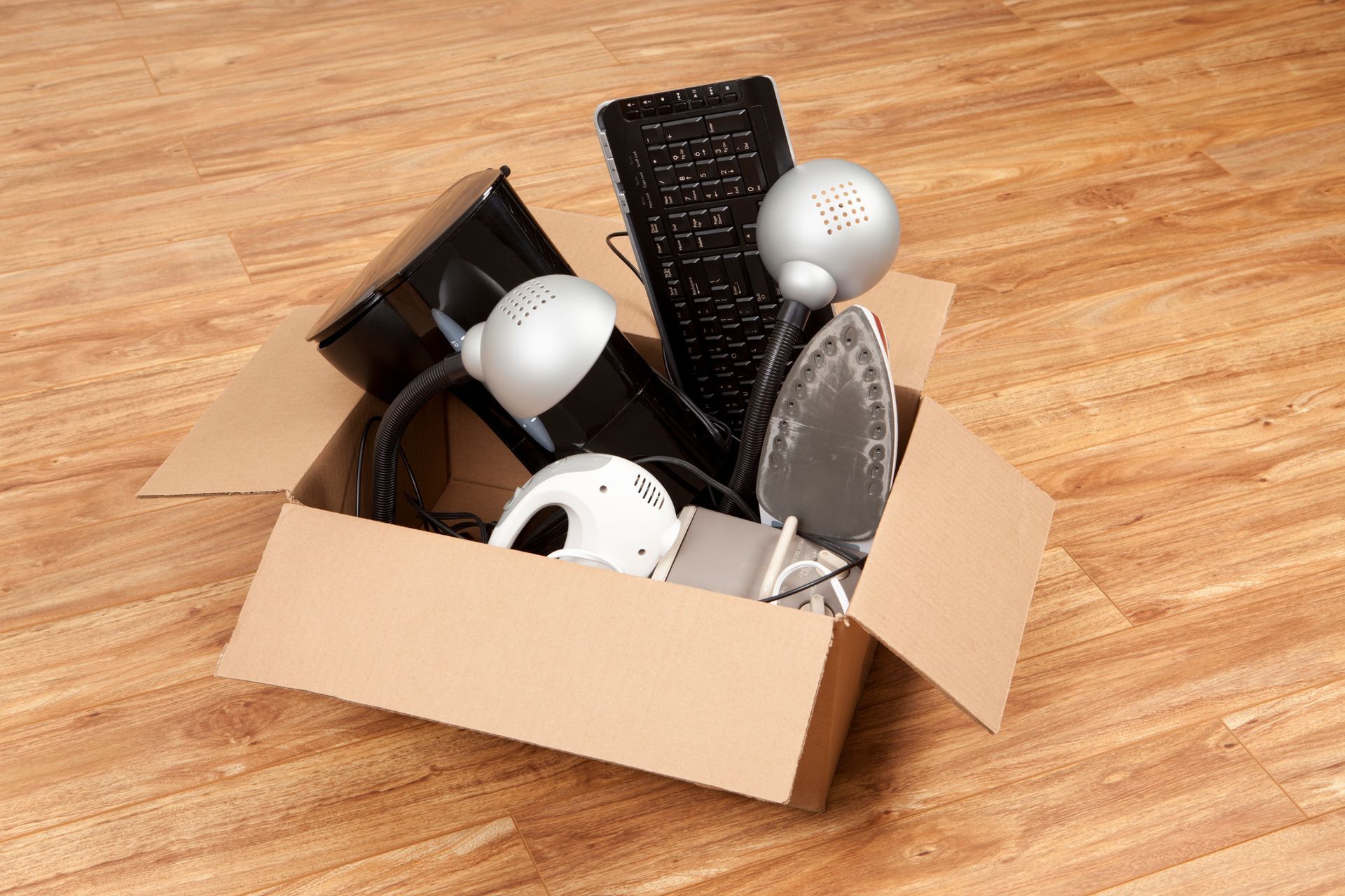 Cardboard box overflowing with various household electronics on a wood floor.