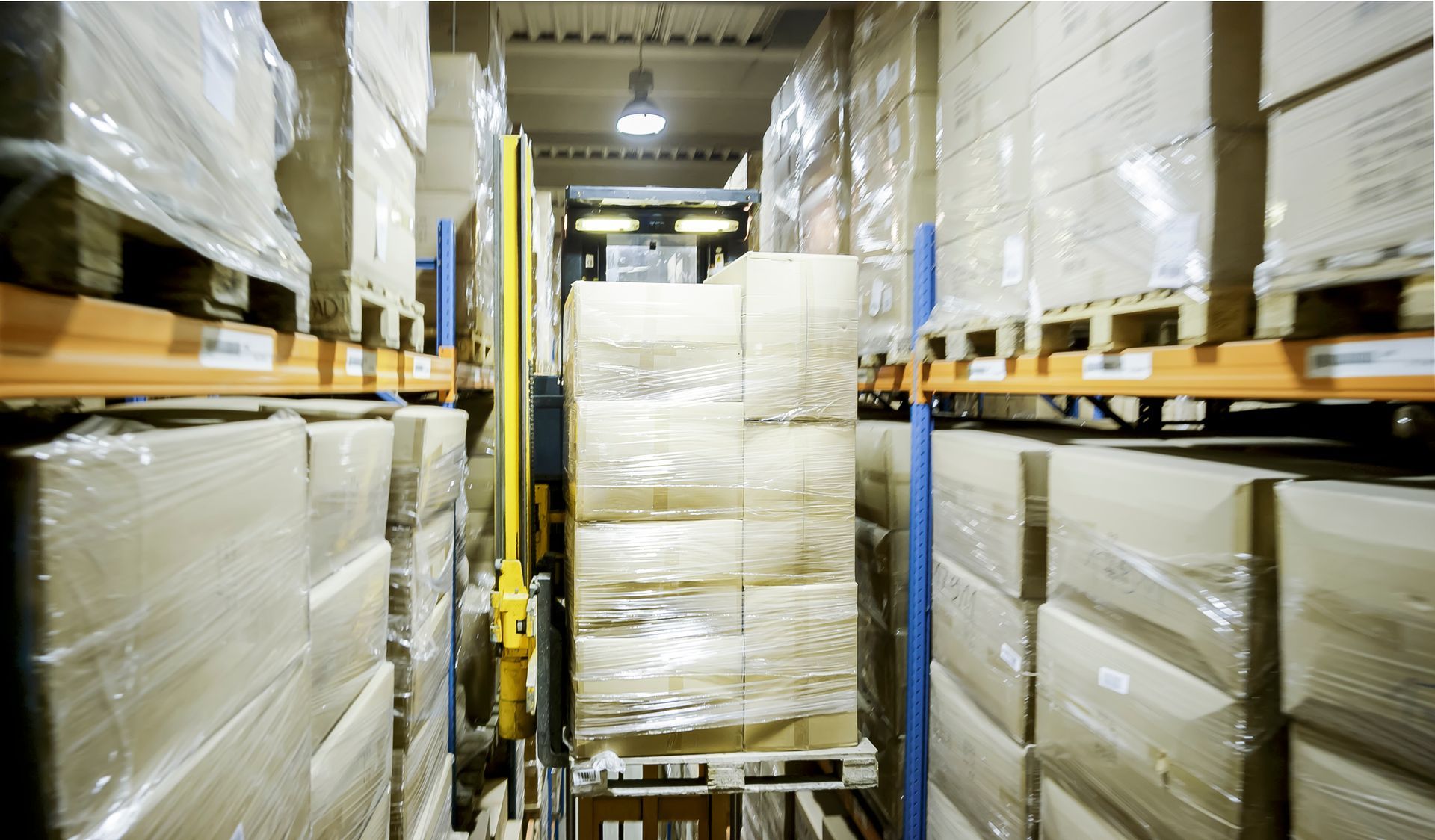 Warehouse aisle with stacked shrink-wrapped pallets on shelves and a pallet jack in the center