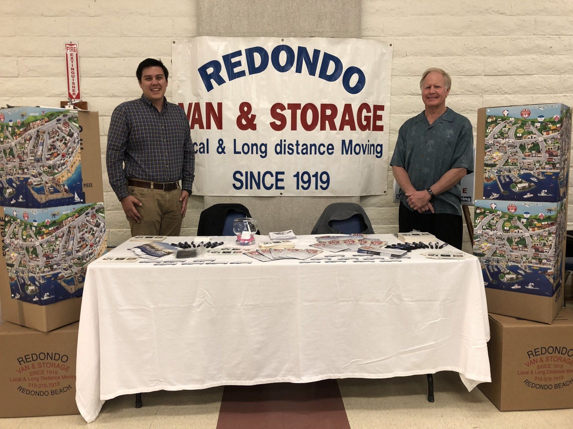 Two men behind a table at a Redon moving and storage booth with banners and stacked boxes