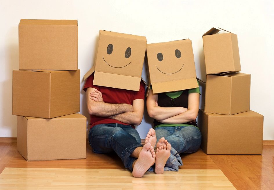 Couple wearing smiley face boxes, leaning against boxes on a hardwood floor, arms crossed.
