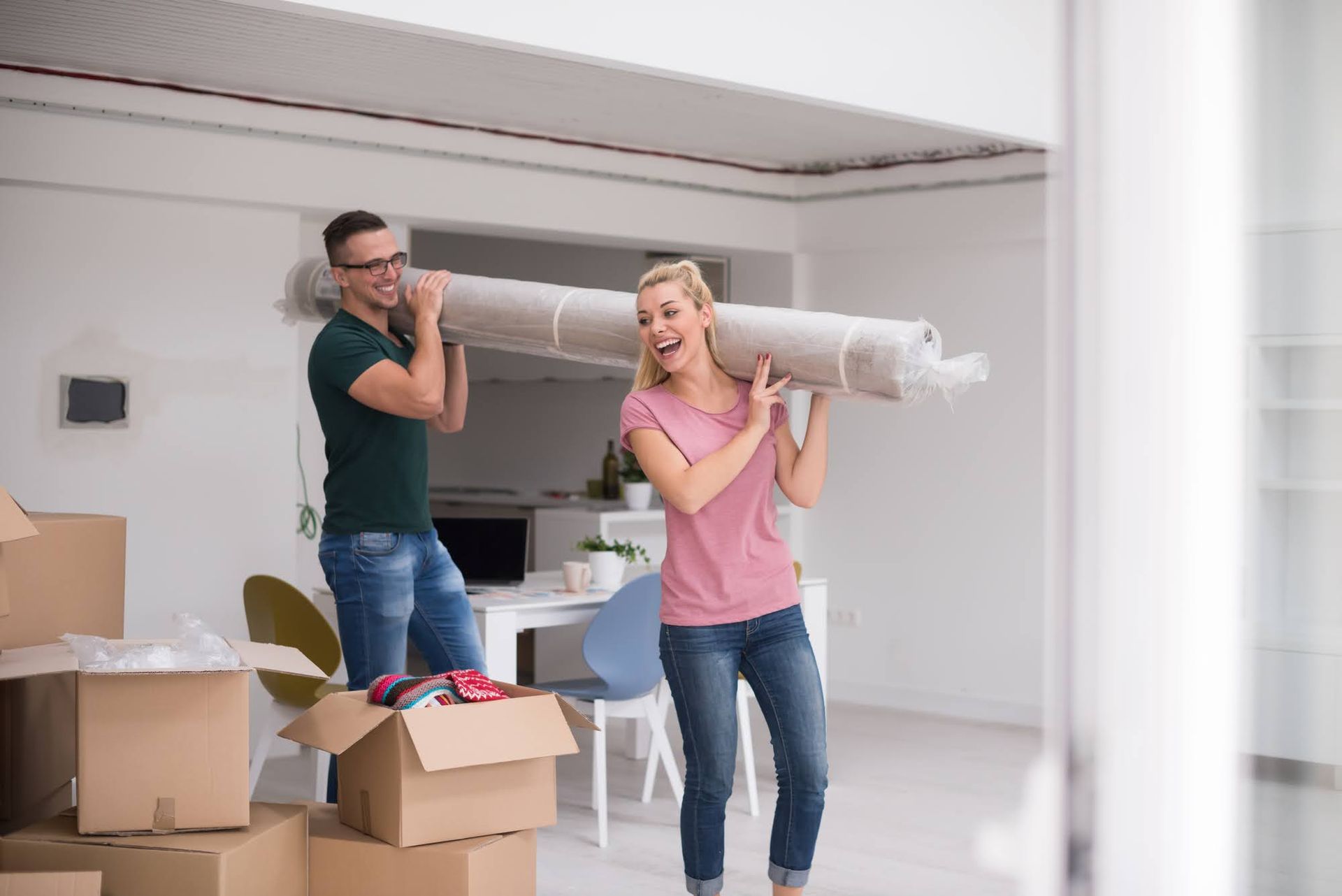 Couple carrying a rolled-up rug indoors, smiling, near moving boxes, in a room with white walls.