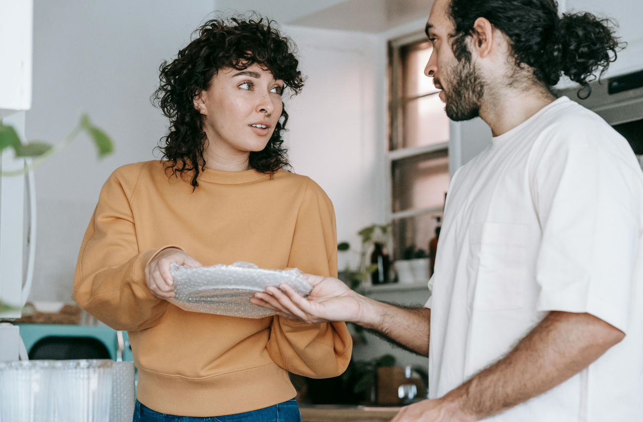 Two people talking in a kitchen while one hands the other a stack of papers
