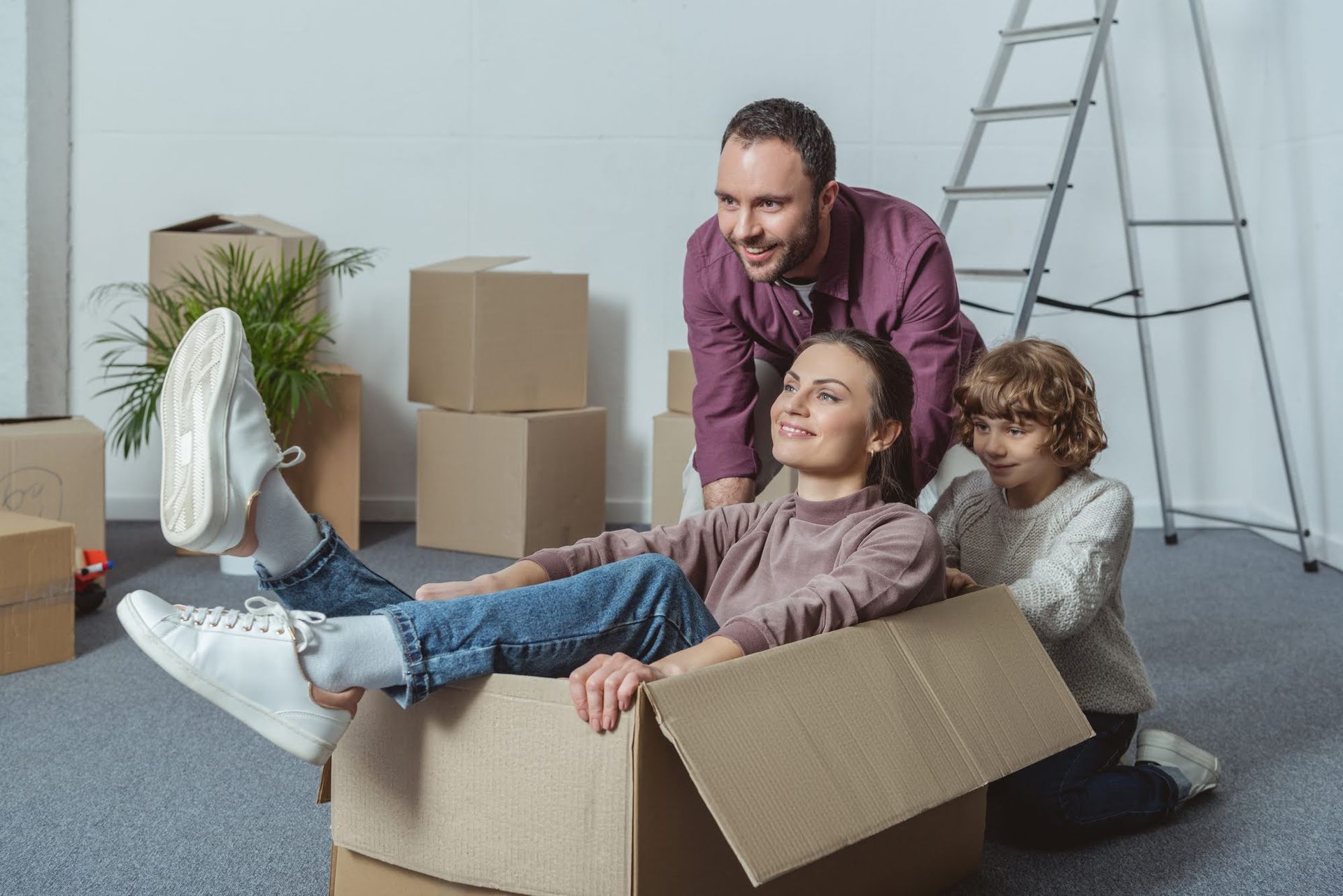Family sitting in a cardboard box in a moving room, smiling among packed boxes and a ladder
