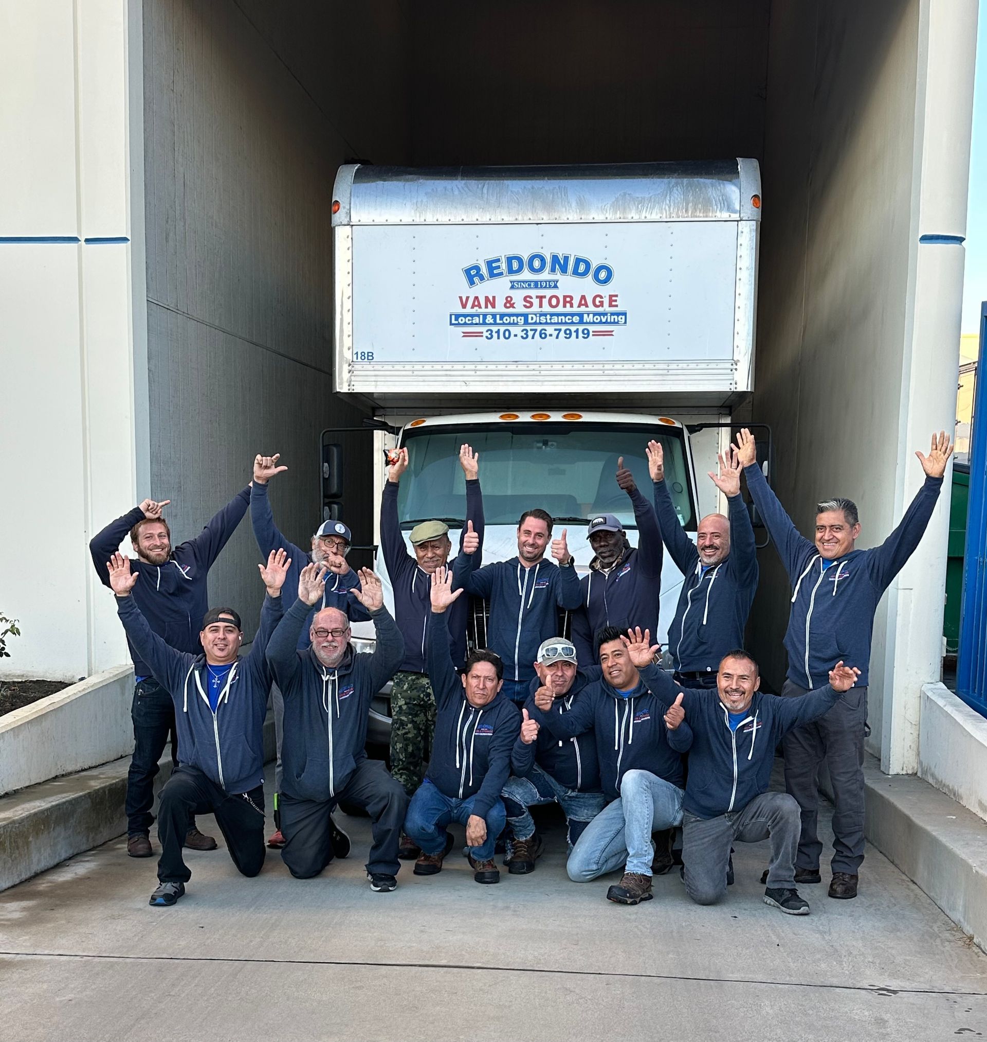 Group of movers posing in front of a Redondo Van & Storage truck, smiling and raising their arms.