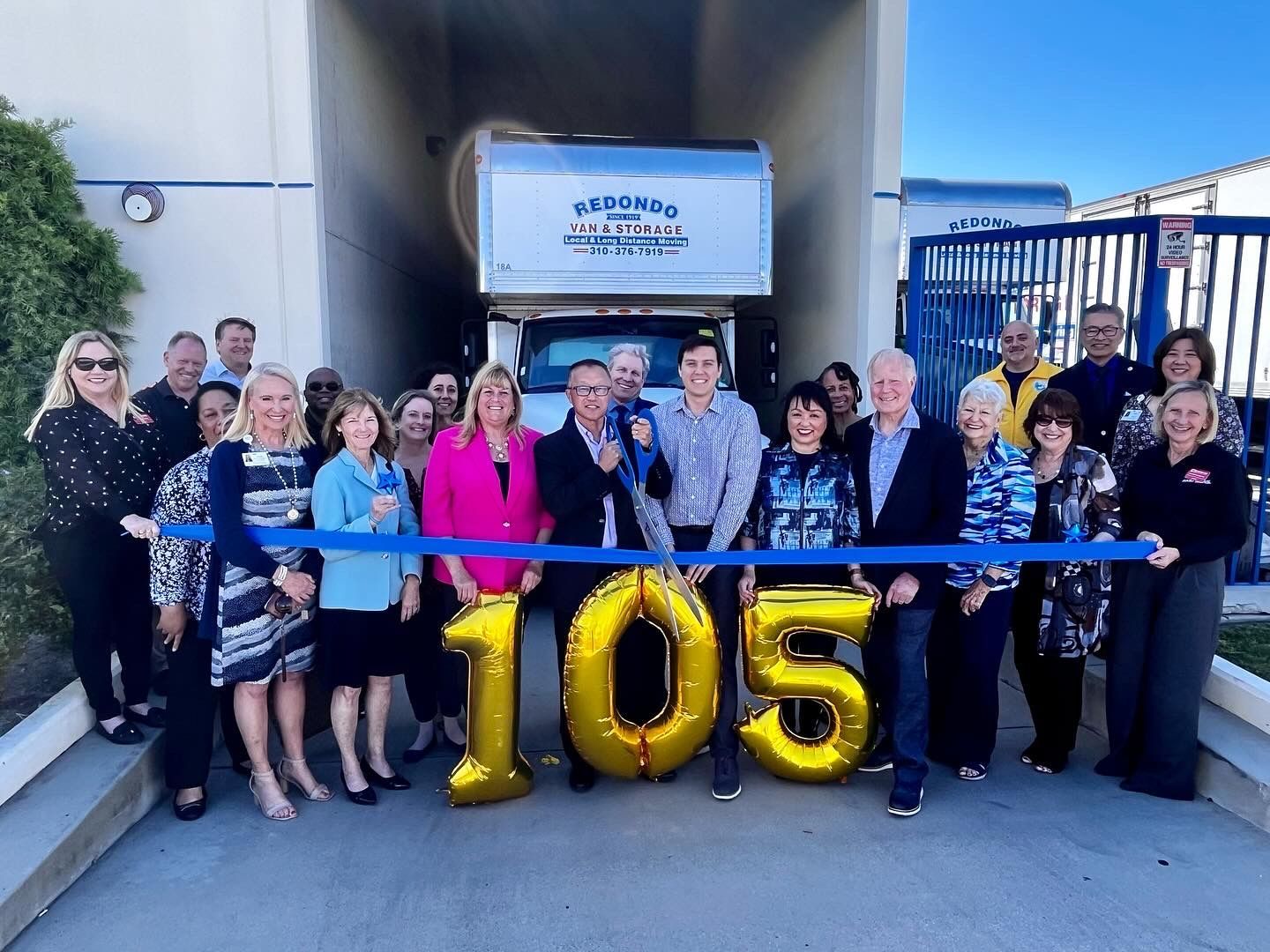 Group of people at a ribbon cutting with large gold “105” balloons outside a building