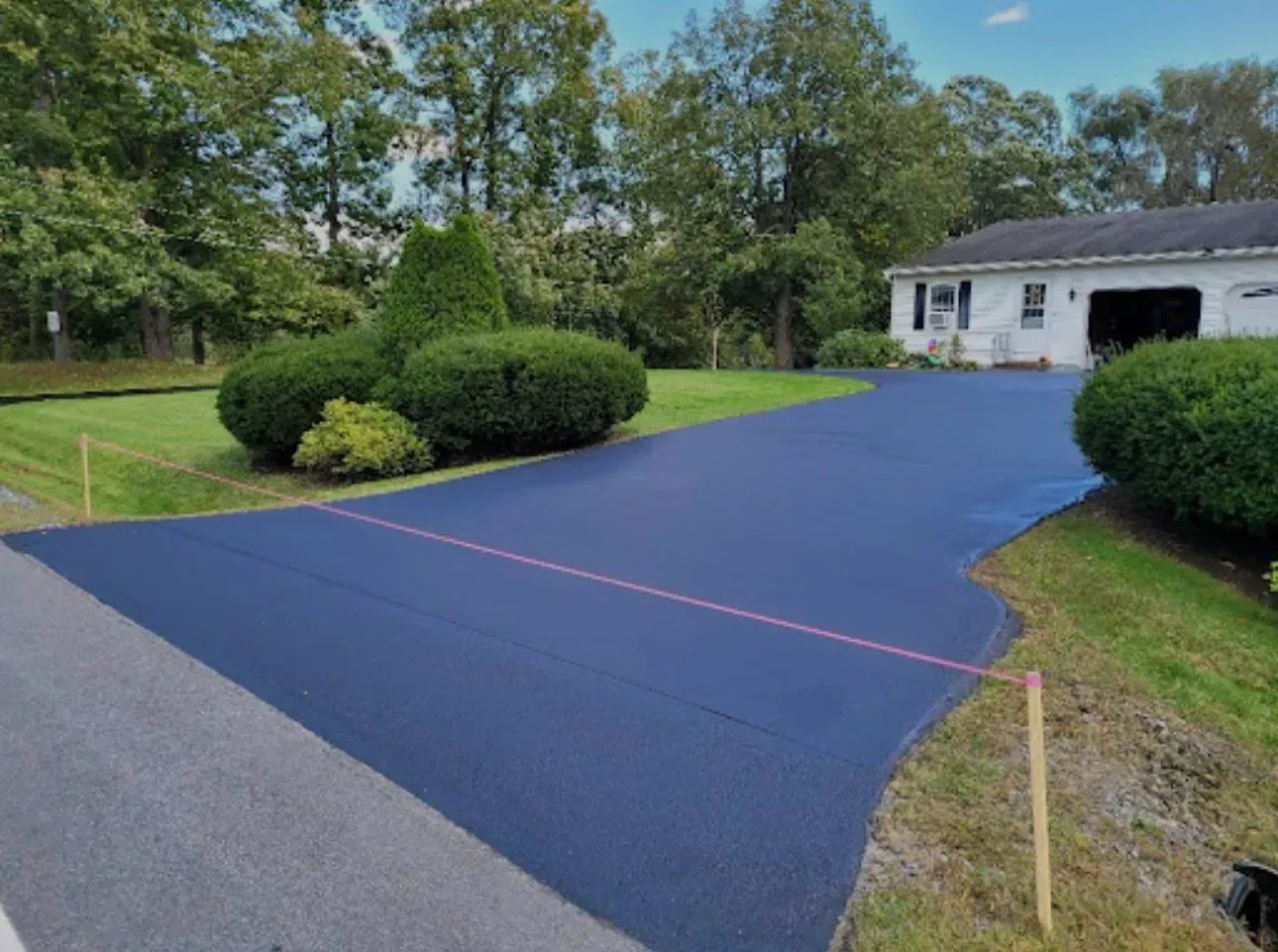 Freshly paved blue driveway beside a white house and green shrubs, with a pink center line and lawn edging
